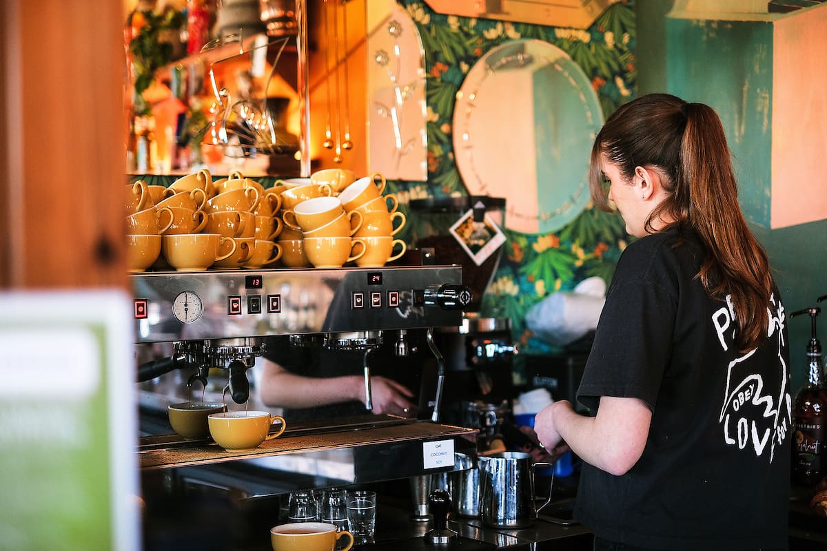 A barista with a ponytail prepares coffee behind a counter, surrounded by stacked yellow rivolo cups and a coffee machine, in a cozy café with colorful, patterned walls.