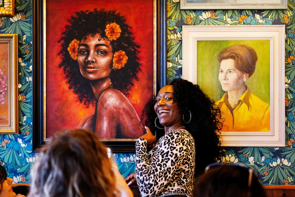A smiling woman with curly hair and glasses poses in front of a colorful rivolo portrait featuring a woman with flowers in her hair, surrounded by other framed portraits on a floral-patterned wall.