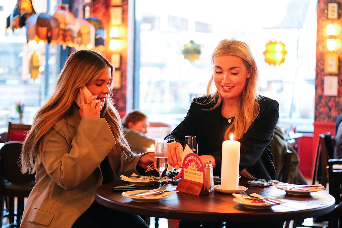 Two young women sit at a round table in a warmly lit restaurant. One checks her phone while the other pours a drink from a bottle of Rivolo. A lit candle, plates, and glasses are on the table, with colorful lights in the background.