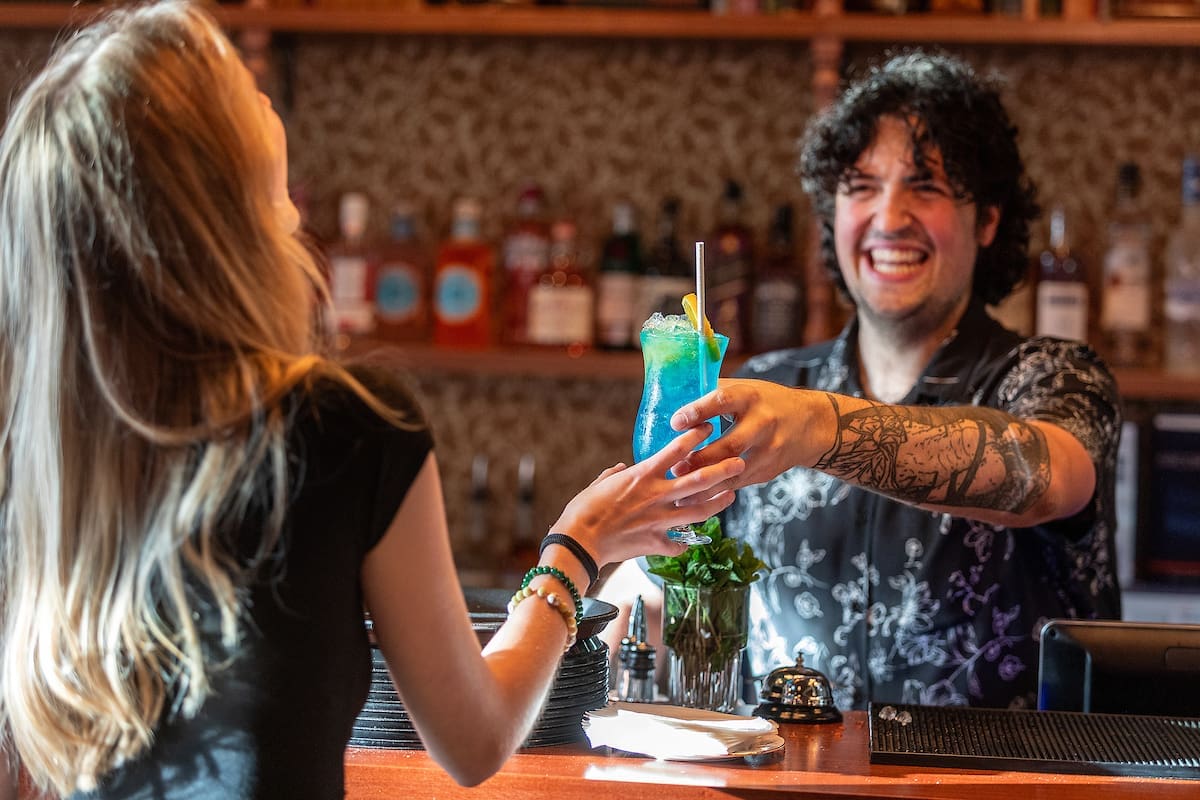 A smiling bartender with tattoos hands a blue cocktail with a straw and garnish to a woman across the lounge bar, with bottles of liquor visible on shelves in the background.