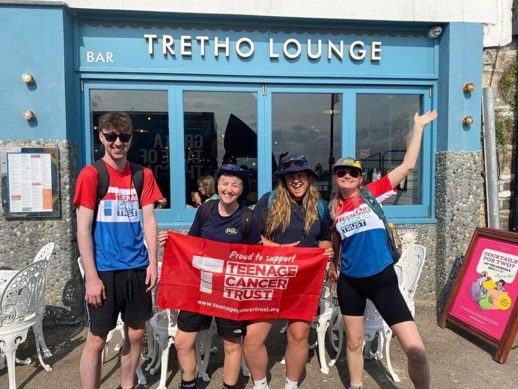 Four smiling people in cycling gear stand outside Tretho Lounge café, holding a red "Teenage Cancer Trust" banner. The group appears happy and energetic, posing with arms raised in celebration—capturing the spirit of our About Us story.