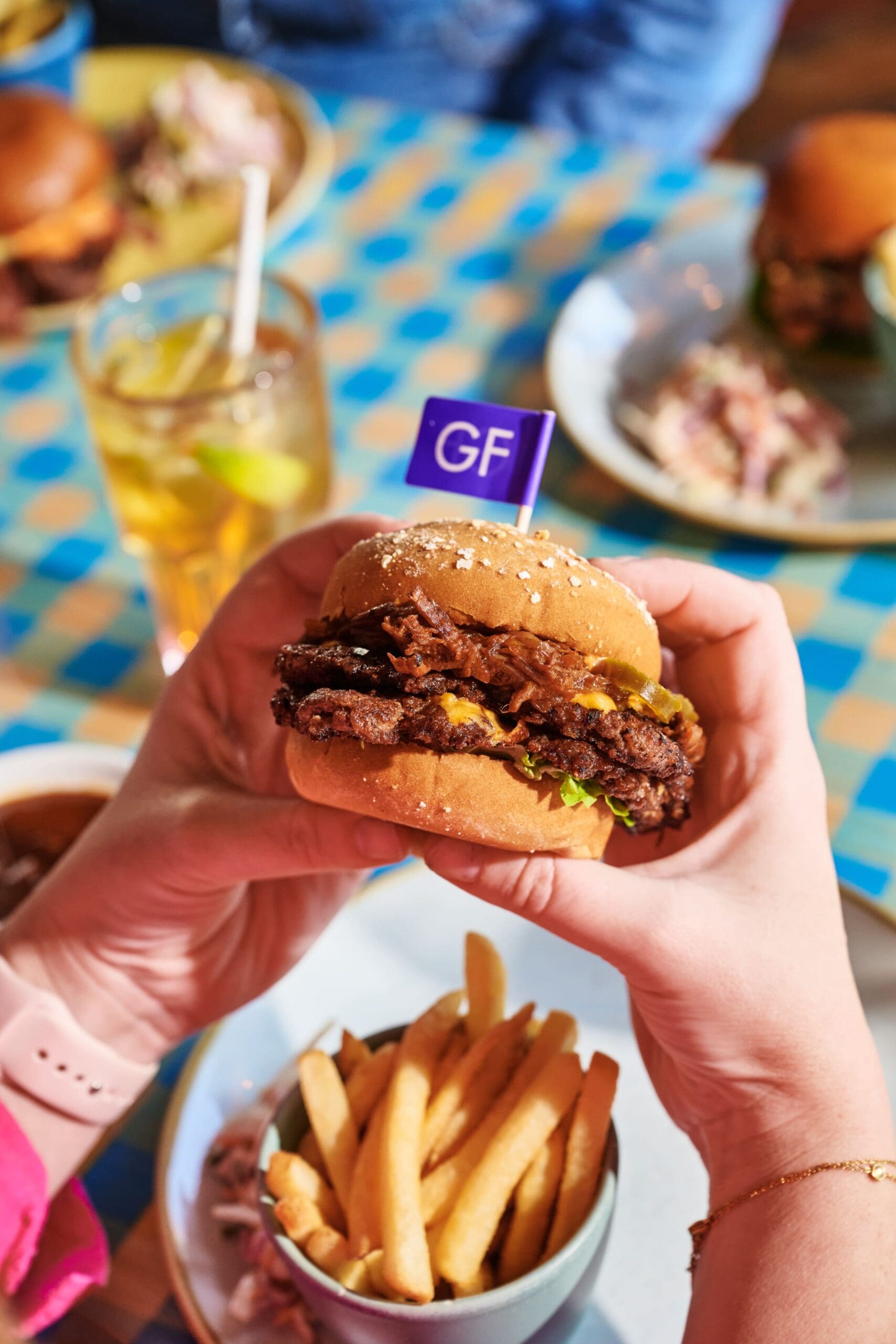A person holds a gluten-free burger with a "GF" flag, above a bowl of fries. The scene includes a drink with lemon and other plates of food on a blue-checkered table.