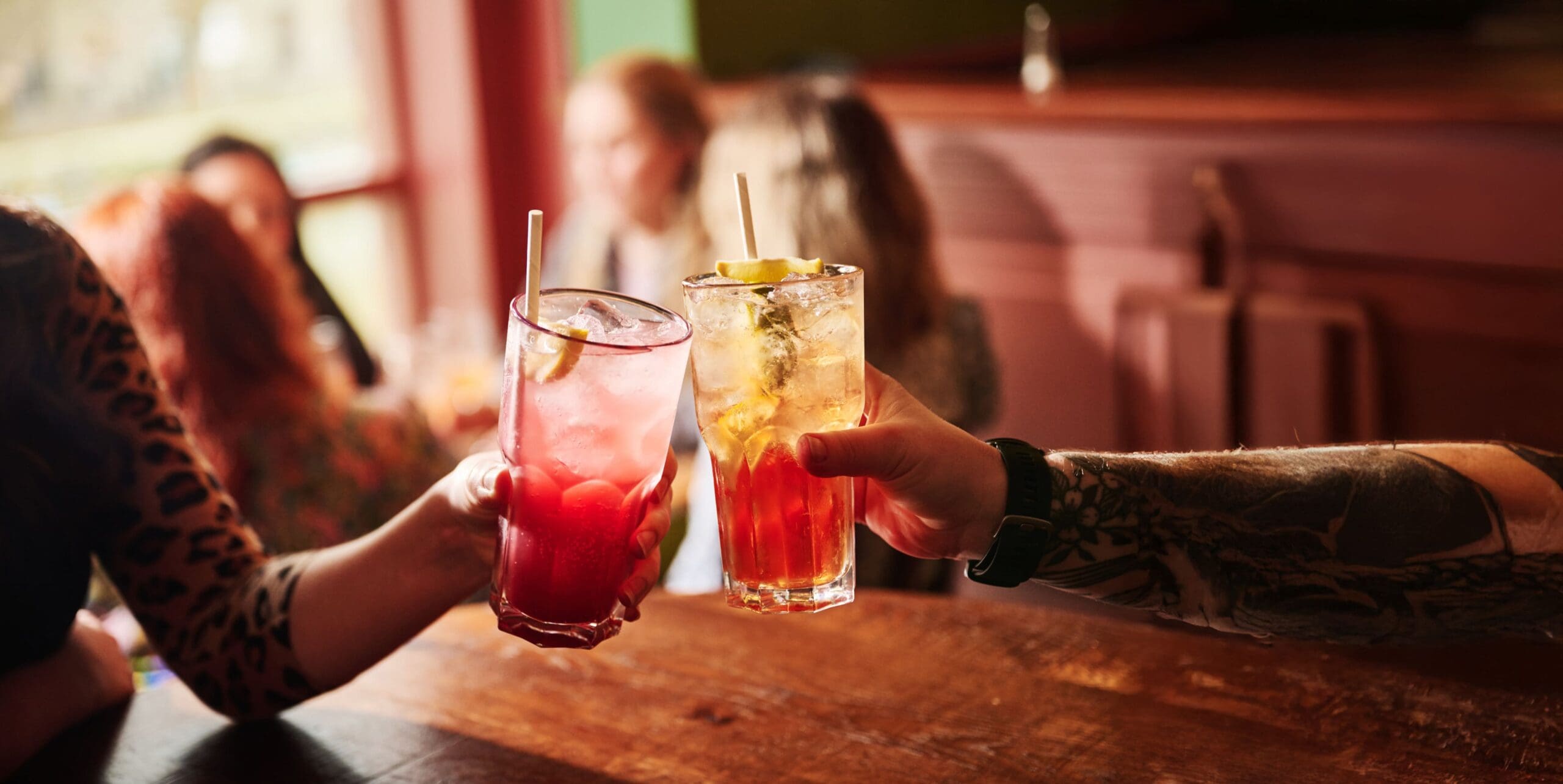 Two people holding up colorful cocktails garnished with lemon slices and straws, toasting over a wooden bar table, with a blurred background of people sitting and talking in a cozy, warmly lit setting.