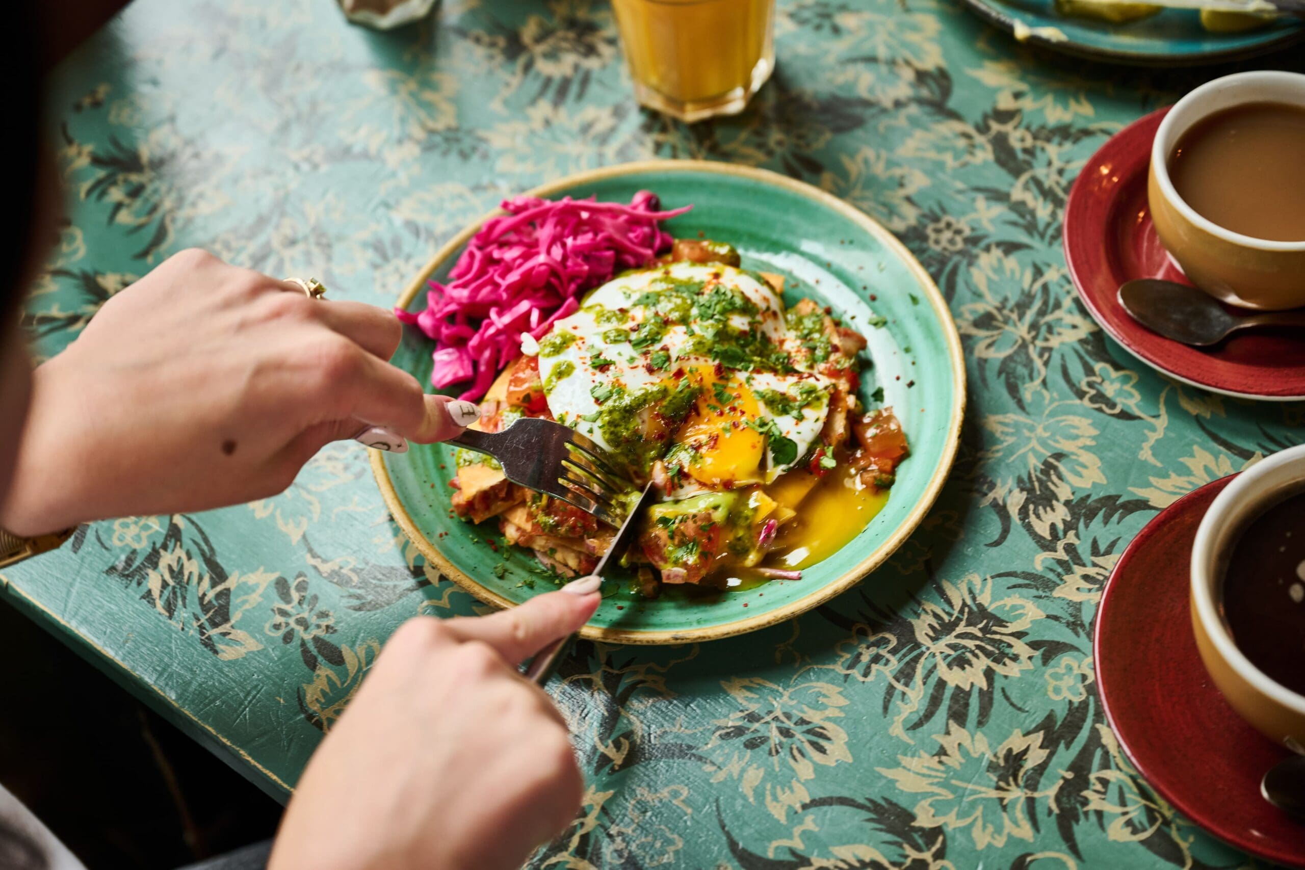 A person cuts into a dish topped with eggs and herbs, served with a side of shredded pink cabbage on a green plate, on a patterned green table next to cups of coffee and a glass of juice.