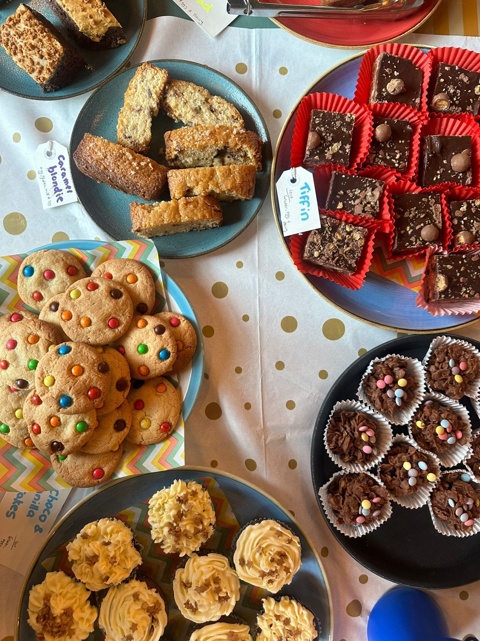 An overhead view of a table with assorted baked treats, including M&M cookies, cupcakes with yellow frosting, chocolate squares in red wrappers, flapjacks, oat bars, and chocolate clusters, all on colorful plates.