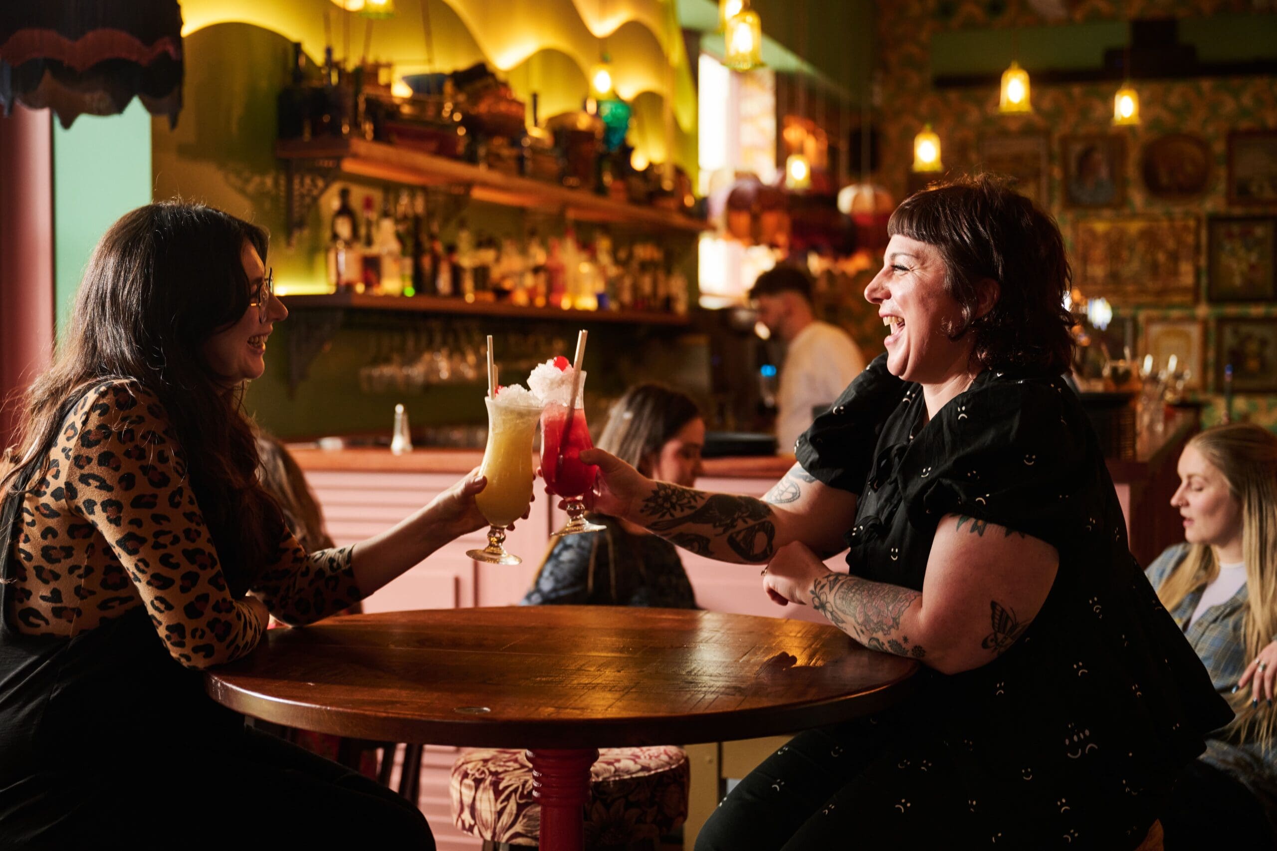 Two women sit at a round table in a lively bar, smiling and clinking colorful cocktails together. The bar is warmly lit, with several people in the background and a well-stocked bar counter.