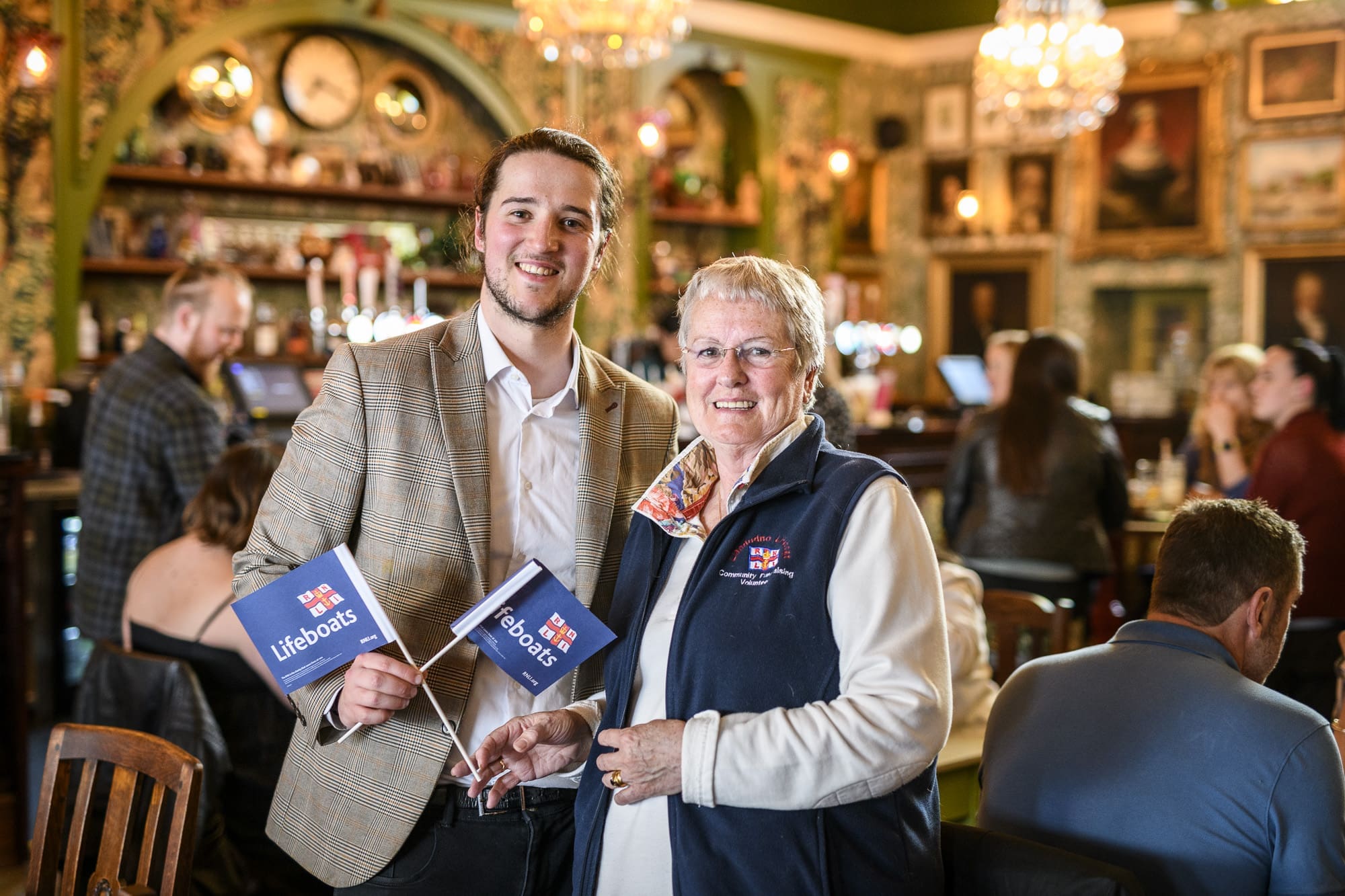 A smiling man and woman stand in a lively, decorated pub holding RNLI Lifeboats flags. The woman wears a navy vest and glasses; the man is in a light blazer. People chat in the background.