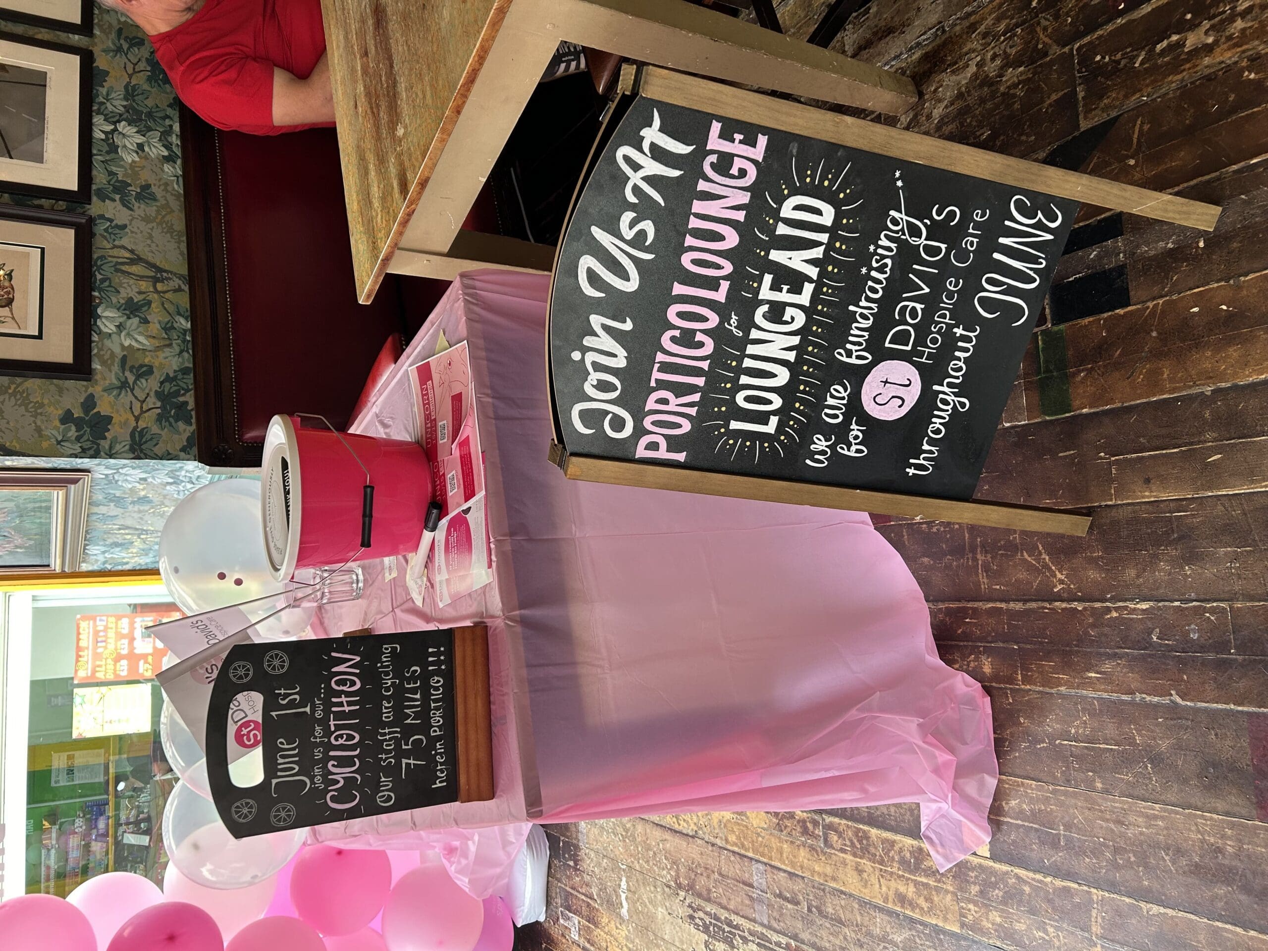A pink-tableclothed fundraising table with donation bucket and two chalkboard signs for a cyclothon event at Portico Lounge, supporting St. David's Hospice Care in June. Balloons and a seated person are nearby.