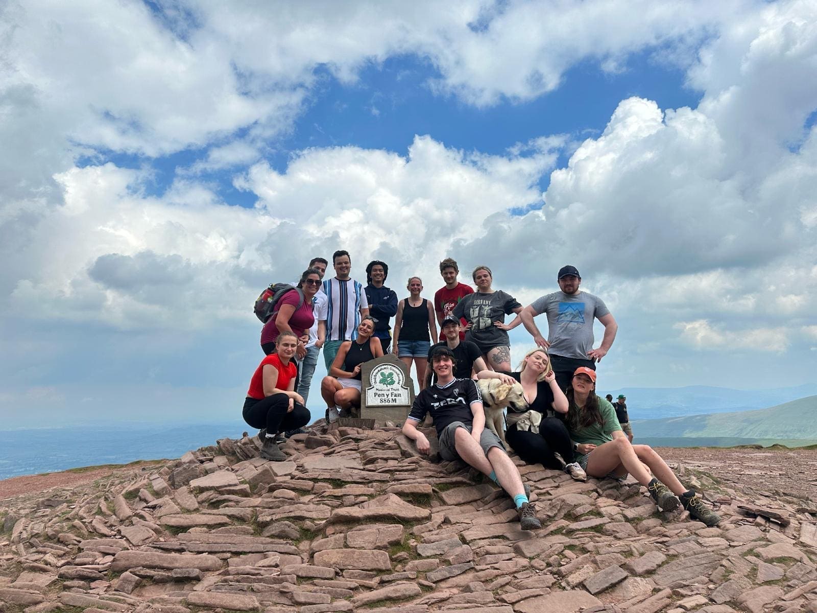 A group of twelve people and a dog pose and smile on a rocky hilltop next to a stone marker under a partly cloudy sky, with distant hills visible in the background.