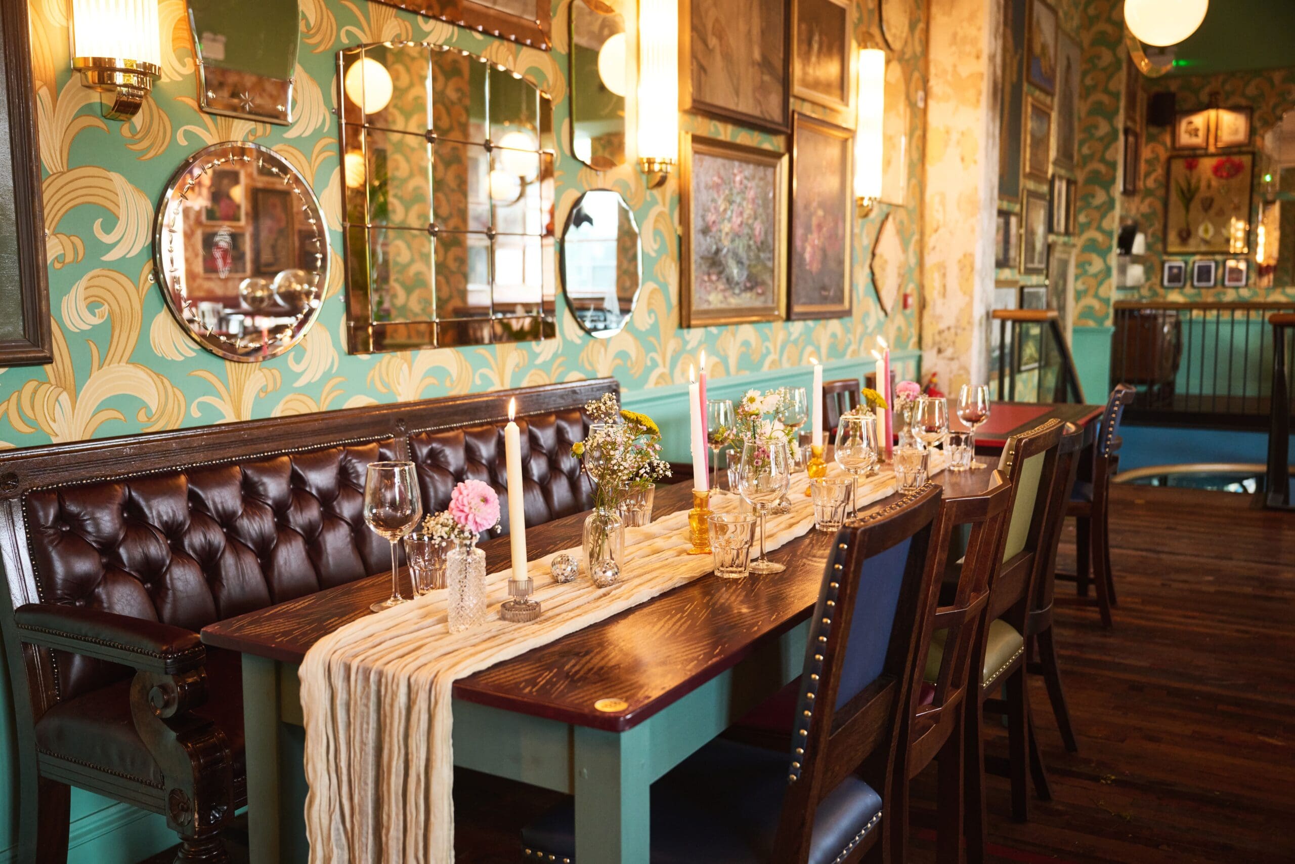 A cozy, vintage-style restaurant with a long wooden table set for dining. The table is decorated with candles, flowers, and glassware. The walls have floral wallpaper, mirrors, and framed art, creating a warm and inviting atmosphere.