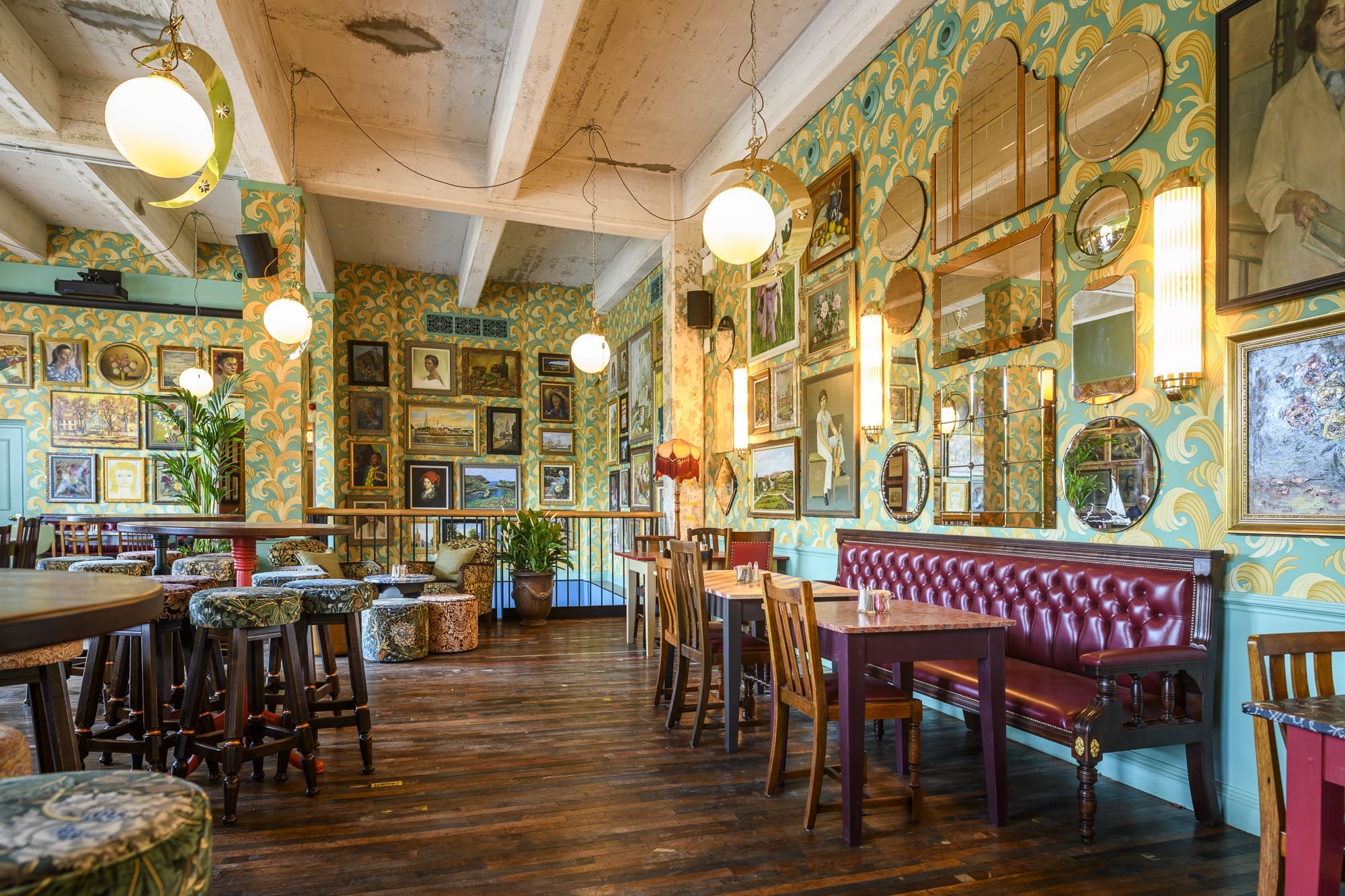 A vibrant, eclectic restaurant interior with patterned wallpaper, framed art and mirrors on the walls, round hanging lights, wooden tables, mixed chairs, and a red leather bench on a polished wooden floor.