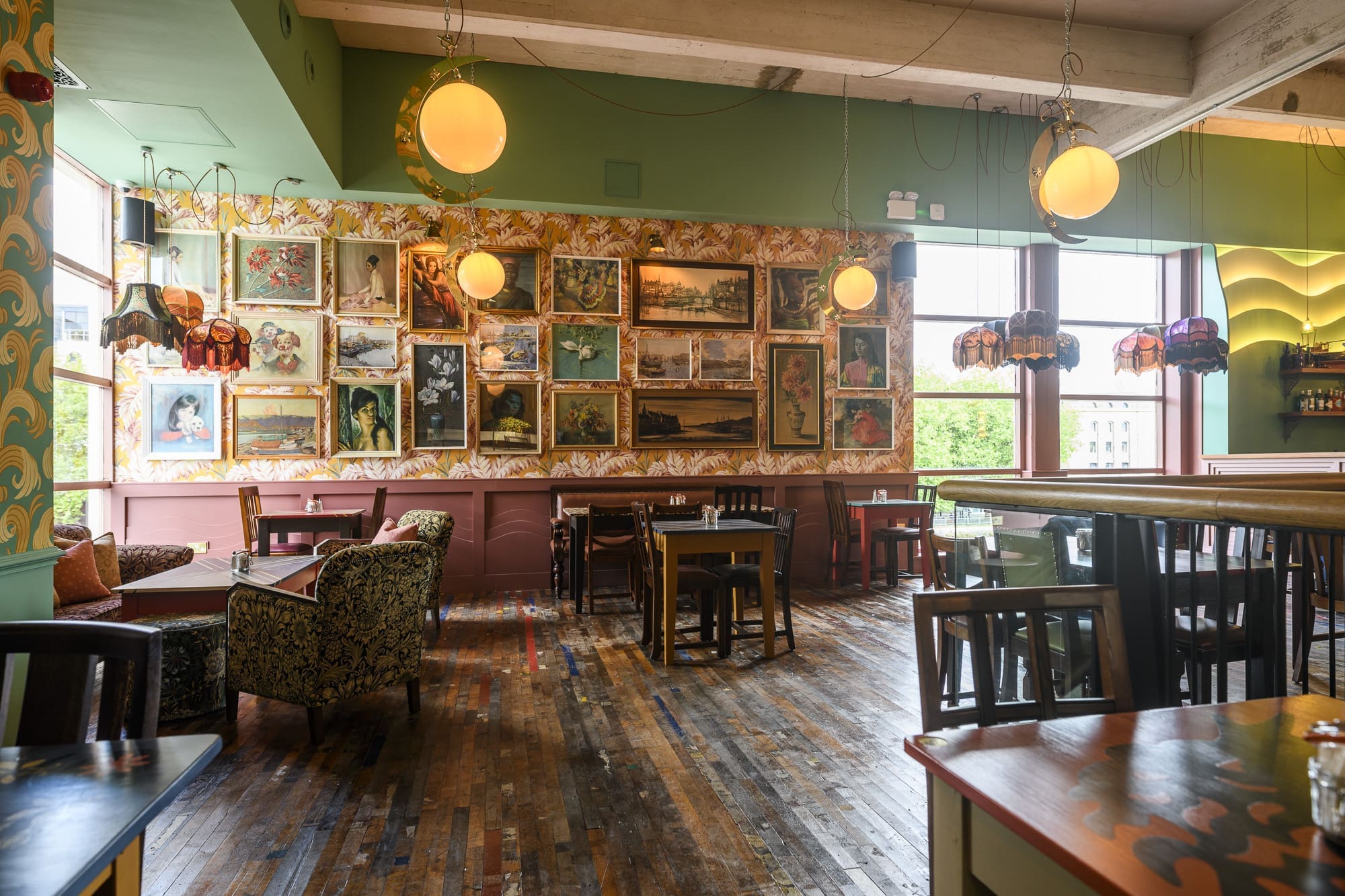 A cozy café with wooden floors, vintage armchairs, round hanging lights, and walls decorated with numerous framed artworks. Large windows let in natural light, and eclectic lamps hang above tables.