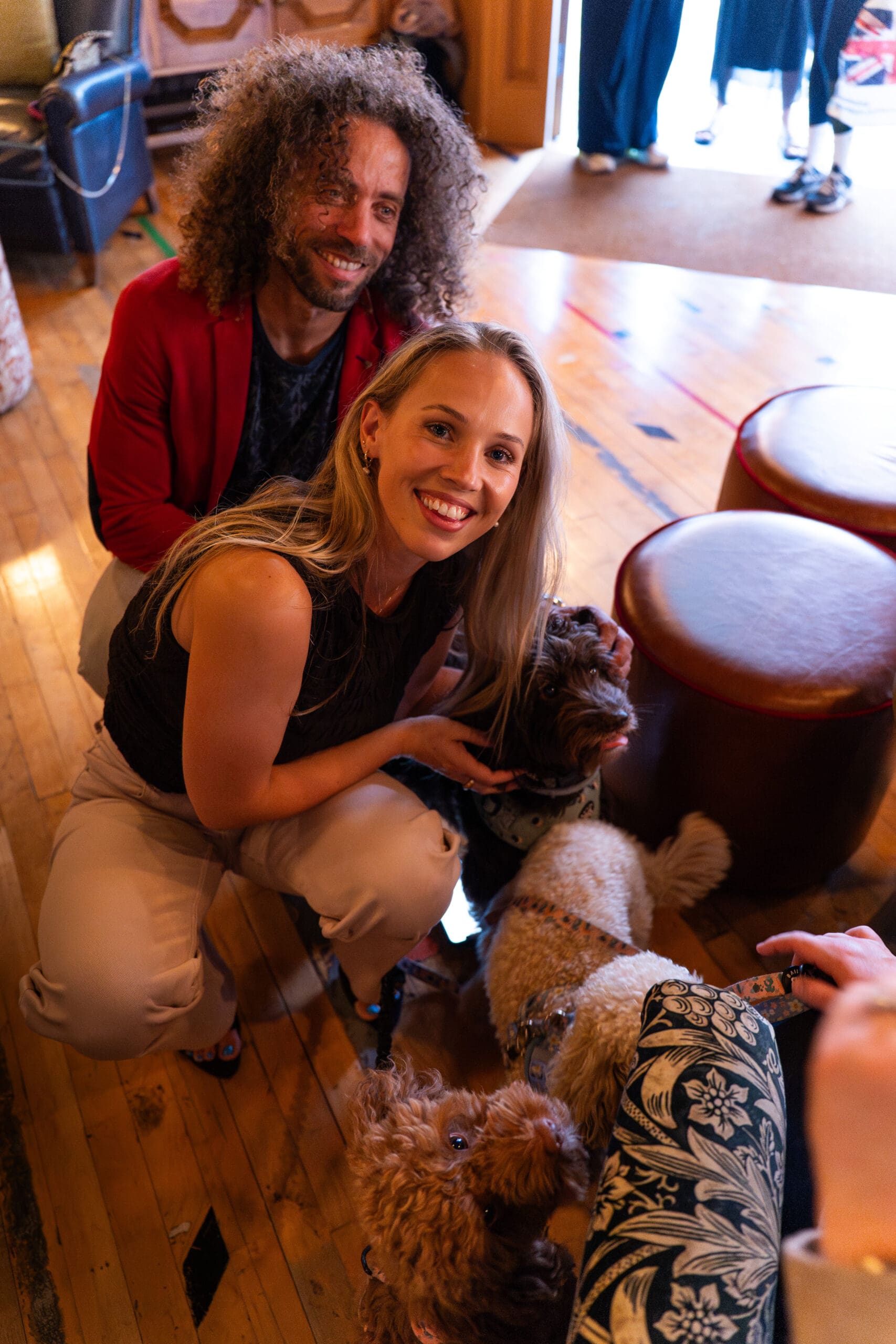 A smiling man and woman kneel indoors, petting two small, fluffy dogs. The woman faces the camera, while the man looks on happily like a cheerful marinero. Warm light fills the cozy room as other people’s legs are visible in the background.