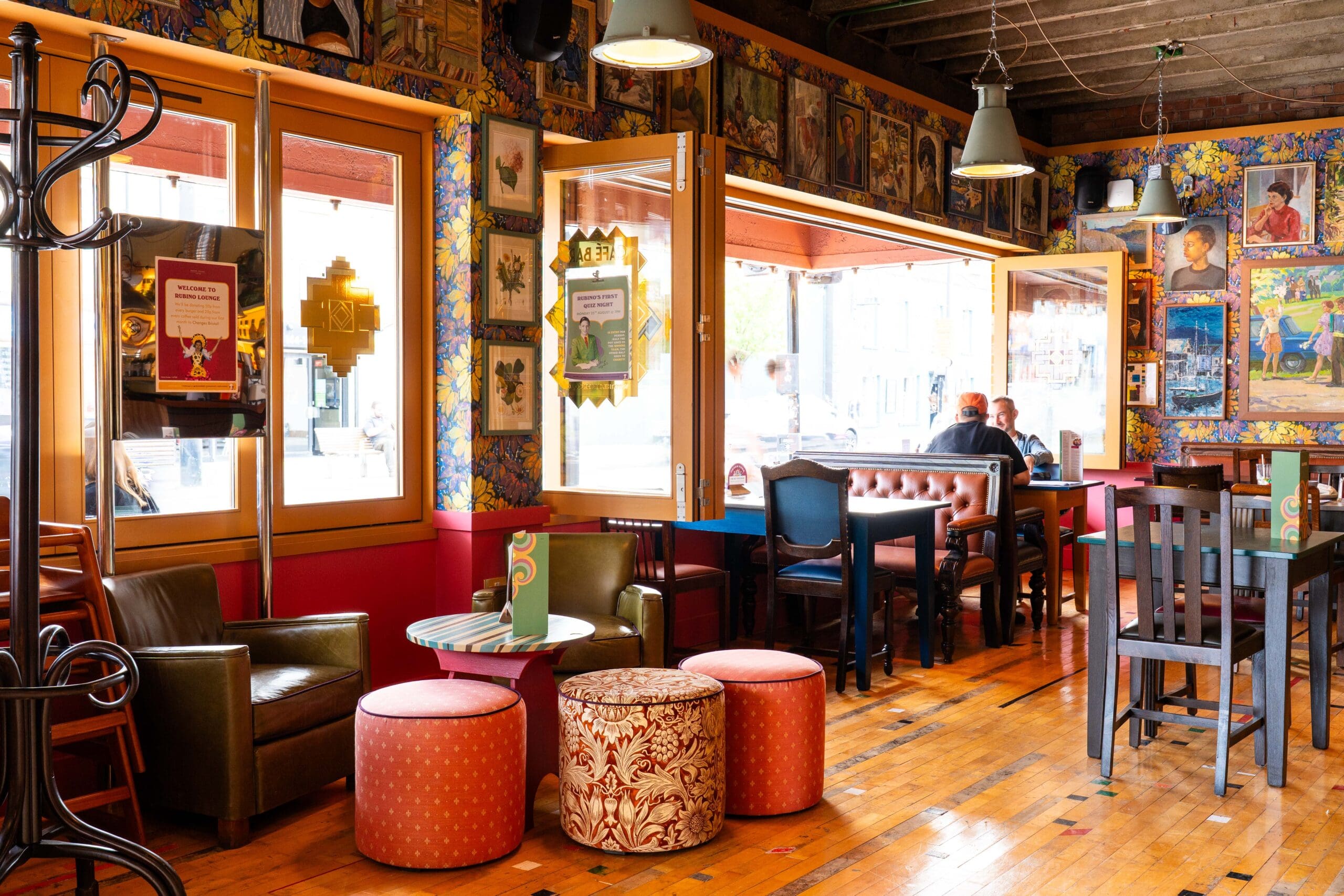 A cozy, colorful cafe with eclectic decor, patterned wallpaper, and framed art. There’s a hint of marinero style in the details, from nautical touches to boat-themed accents. Large windows fill the space with natural light as people chat by the window.