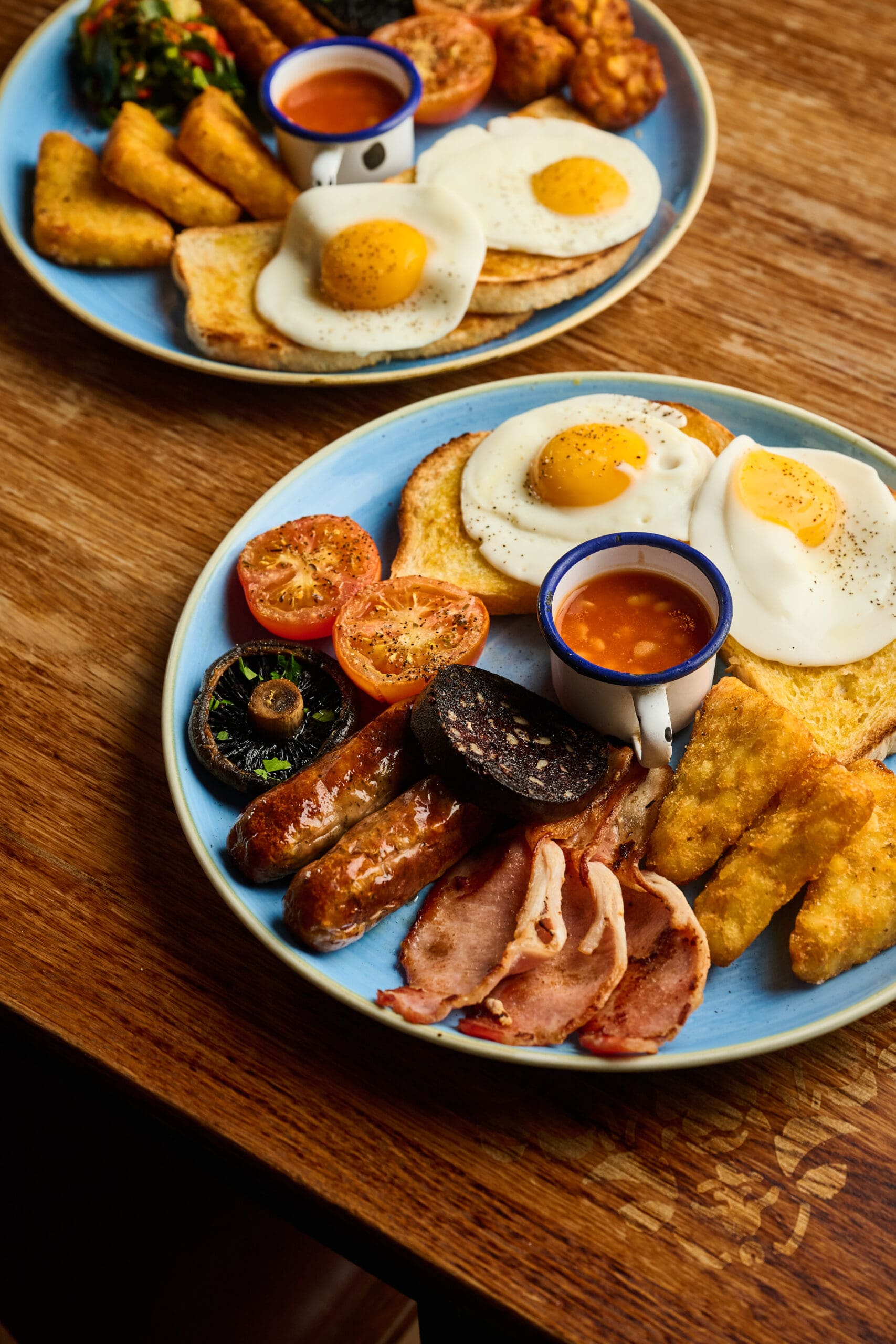 Two blue plates with English breakfasts, including fried eggs, toast, grilled tomatoes, mushrooms, sausages, bacon, black pudding, hash browns, and a small cup of baked beans, set on a wooden table.