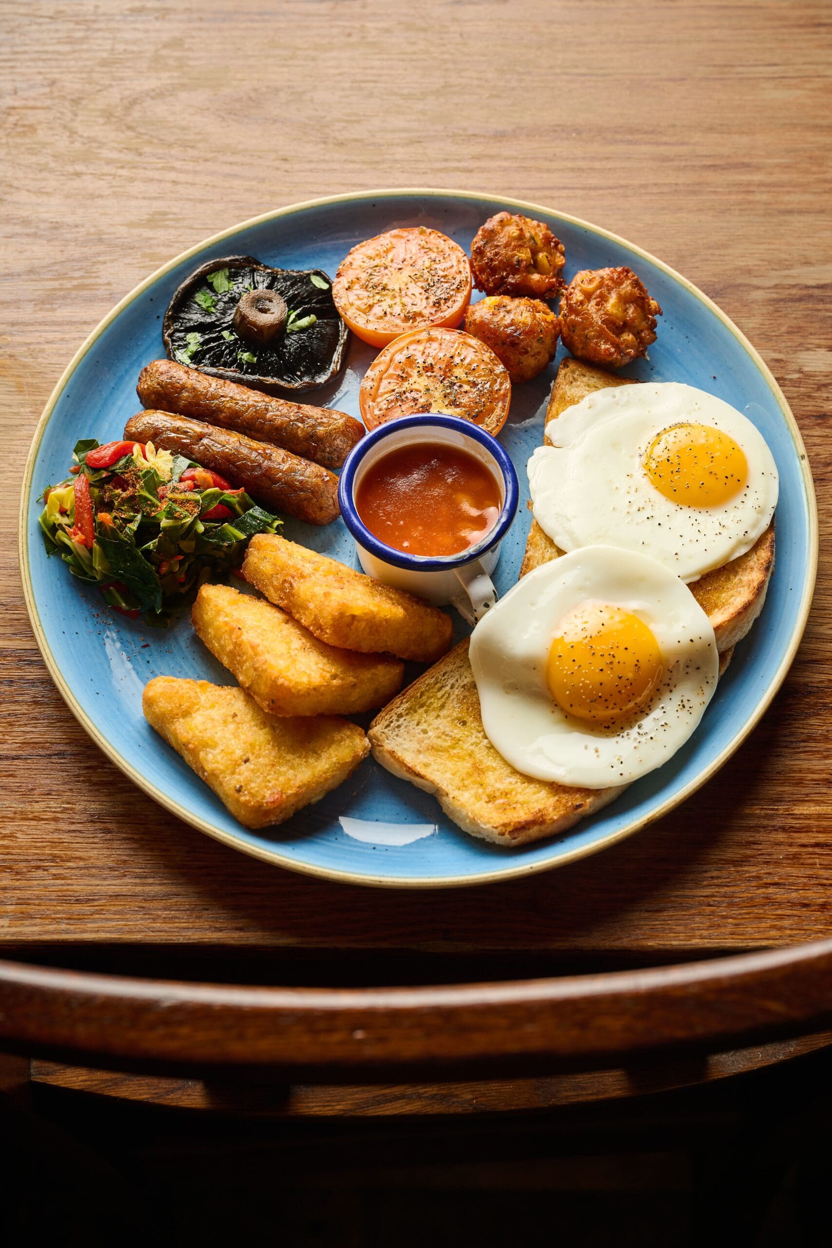 A plate of breakfast food including fried eggs on toast, hash browns, grilled tomato slices, baked beans in a small bowl, sausages, a grilled mushroom, and sautéed vegetables on a blue plate on a wooden table.