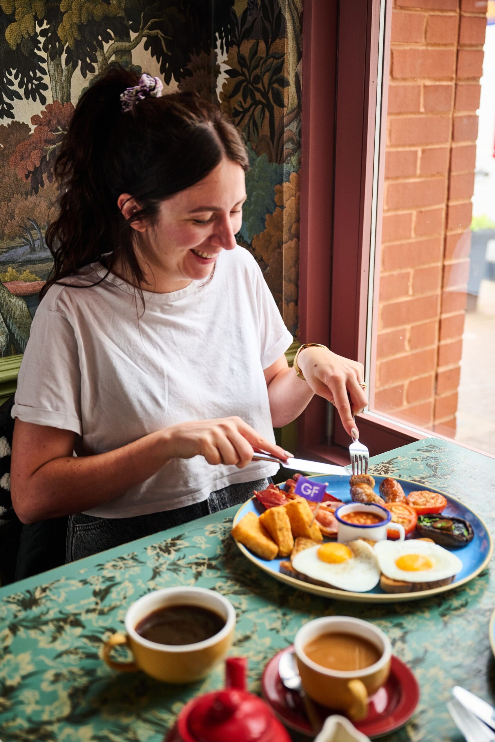 A smiling woman in a white shirt sits at a table with a colorful breakfast plate of eggs, hash browns, beans, sausage, and tomatoes, with coffee and tea cups in front of her by a window.