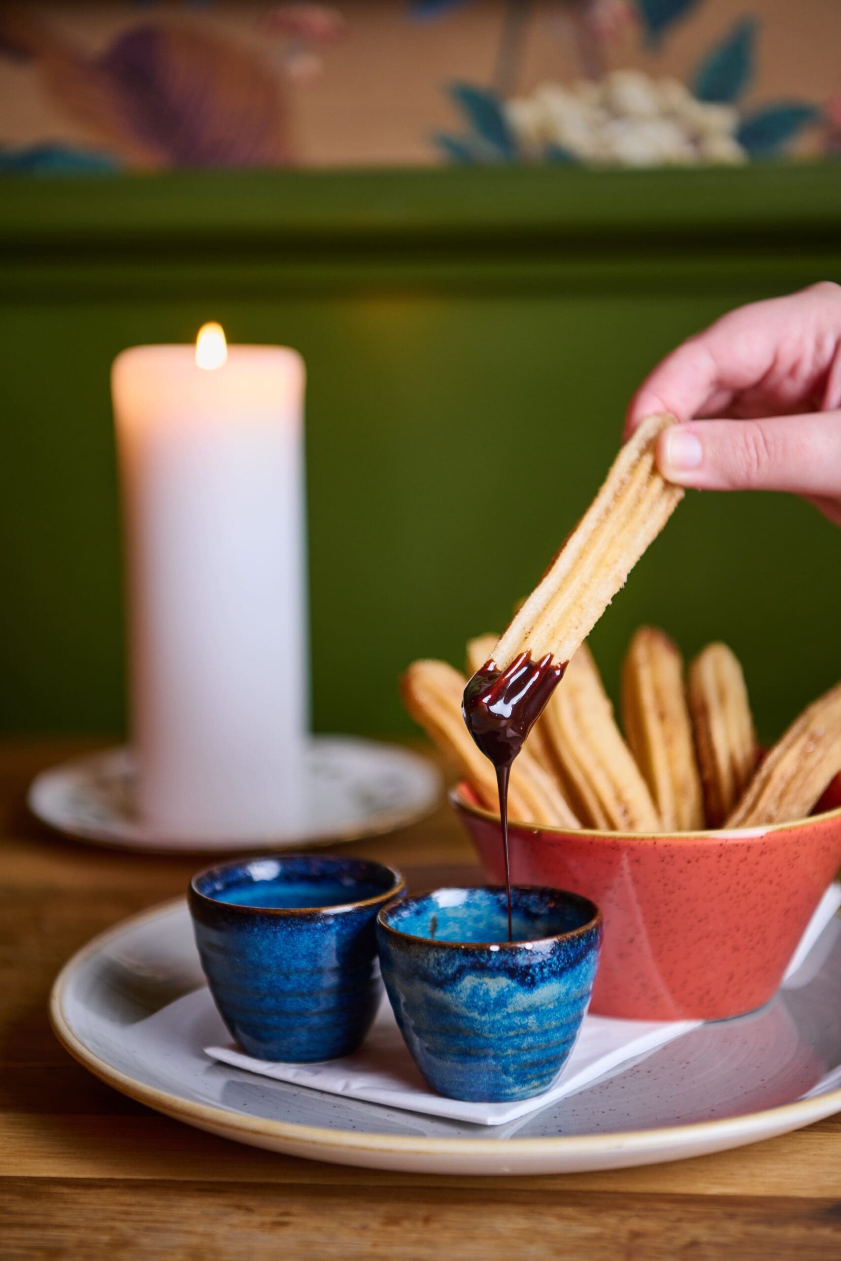 A hand dips a churro into chocolate sauce, with a bowl of churros and two small blue cups on a plate. A lit white candle is in the background on a wooden table.