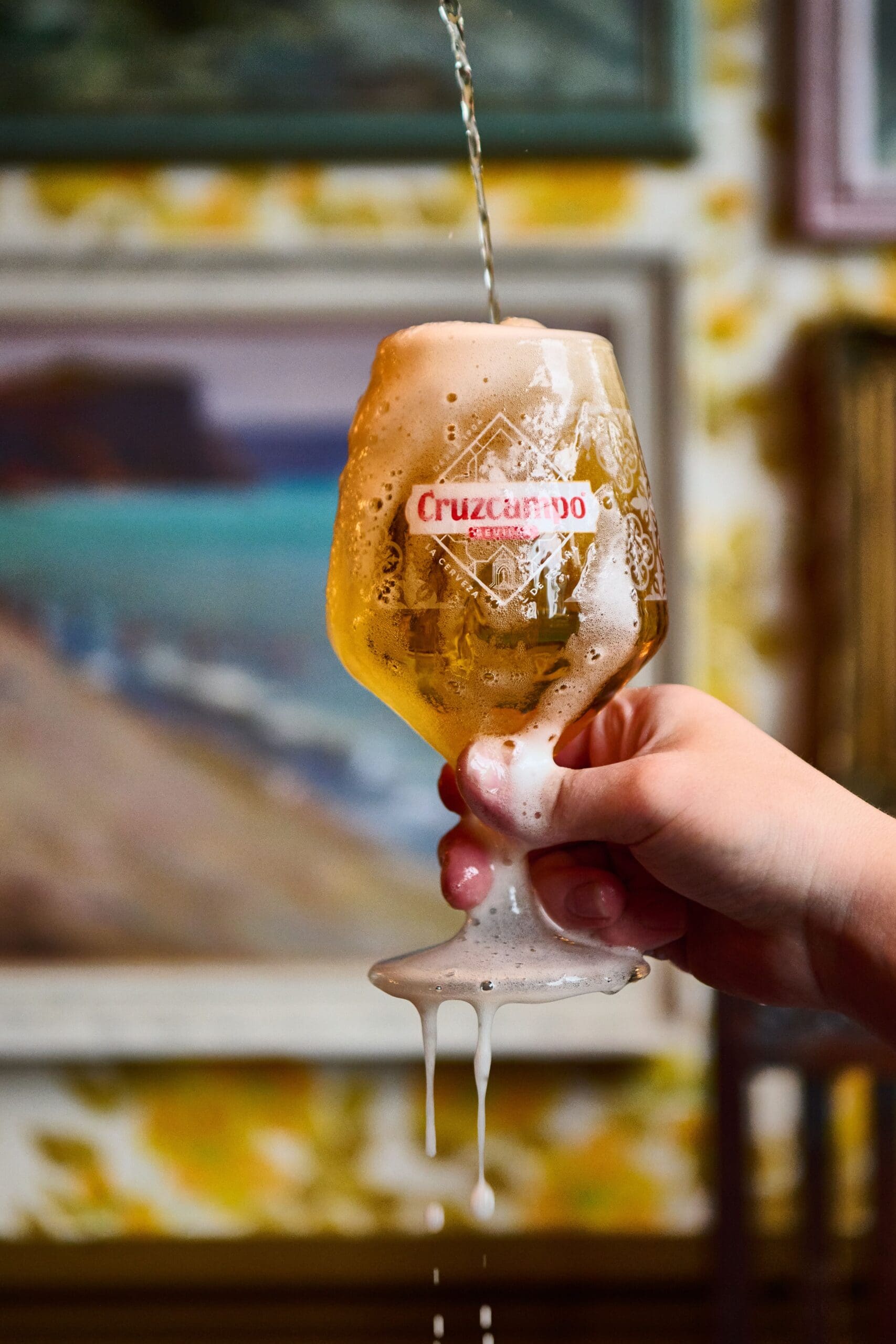 A hand holds a glass of Cruzcampo beer being poured, overflowing with foam and dripping down the sides. Blurry paintings and a patterned wall are visible in the background.