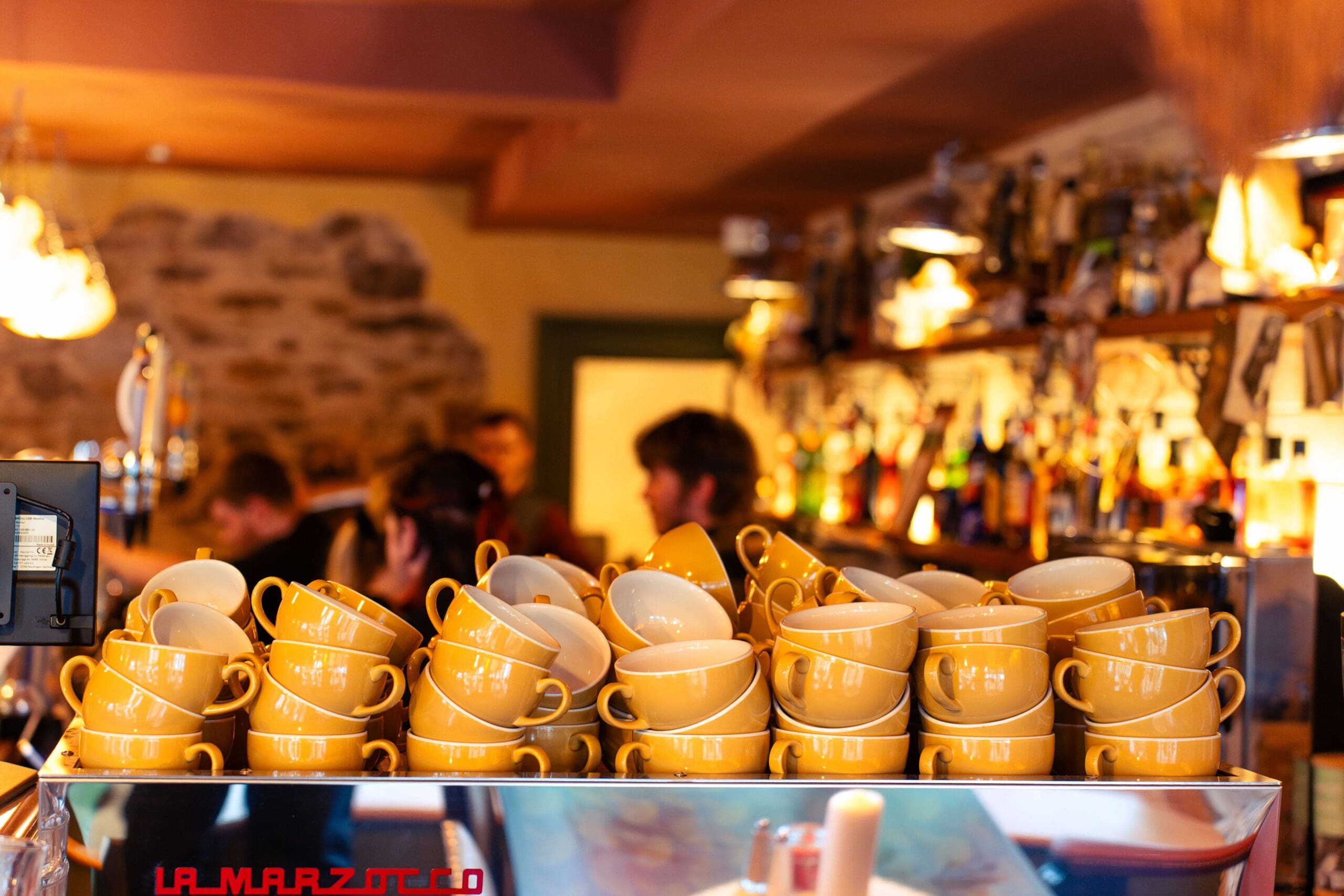 A stack of yellow coffee cups sits on an espresso machine in a cozy café lounge, with a blurred background of people and shelves filled with bottles and glasses.