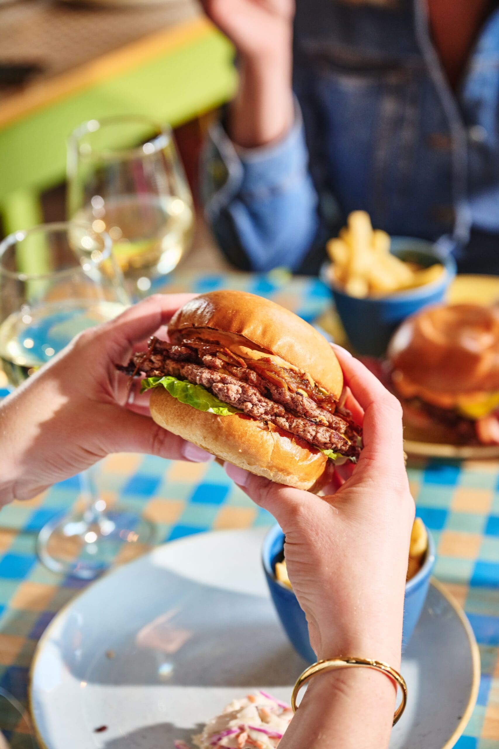 A person holding a stacked beef burger with lettuce in a bun over a plate, with fries, coleslaw, and a glass of white wine on a checkered table. Another person sits across the table.