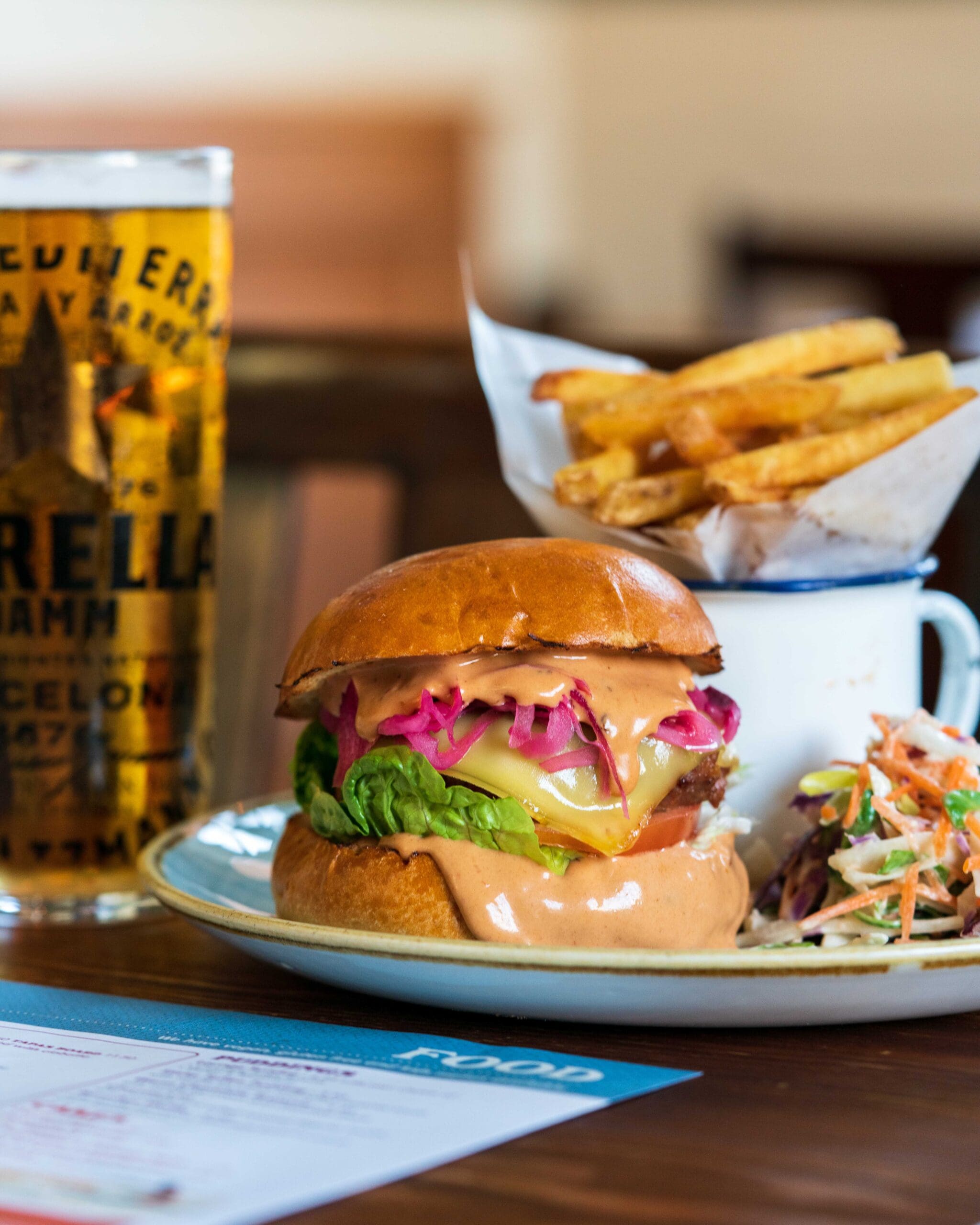 A cheeseburger with lettuce, tomato, and pink slaw, topped with sauce, served with a side of coleslaw and fries in a cup. A tall glass of beer is in the background on a wooden table.