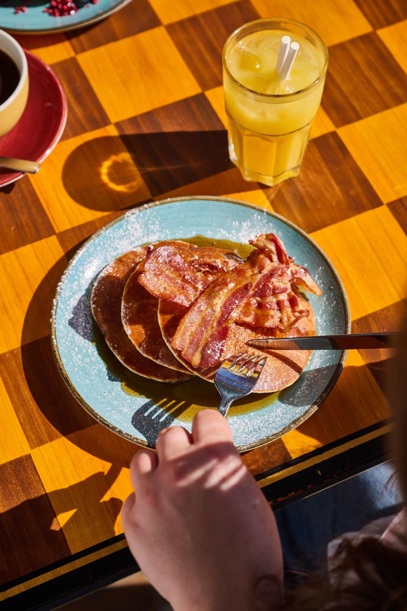 A person lounges while using a fork and knife to eat pancakes topped with bacon on a teal plate, with a glass of orange juice and a coffee cup on a checkered wooden table in bright sunlight.