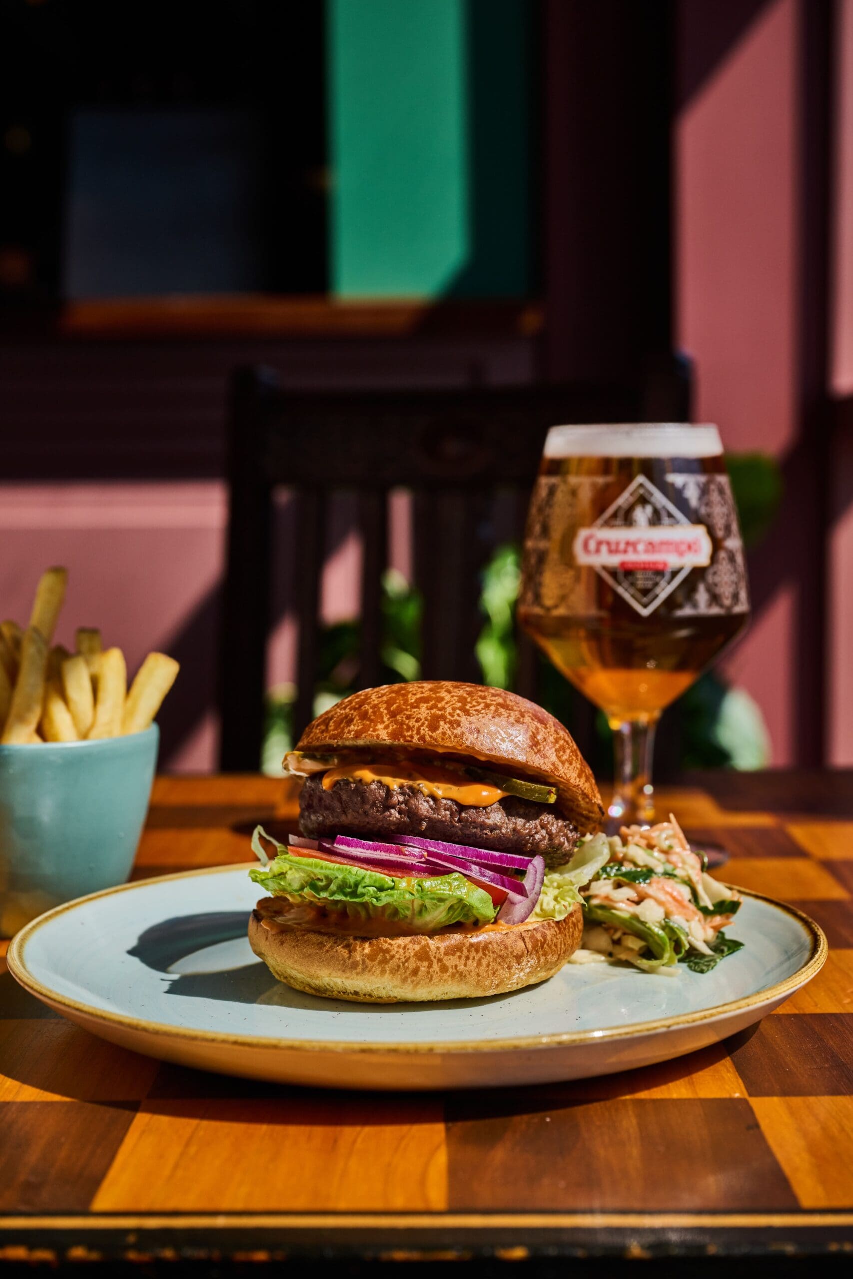A plate with a burger topped with lettuce and red onion sits on a wooden table in the lounge, next to a bowl of fries and a glass of Cruzcampo beer. The background is softly lit by sunlight.