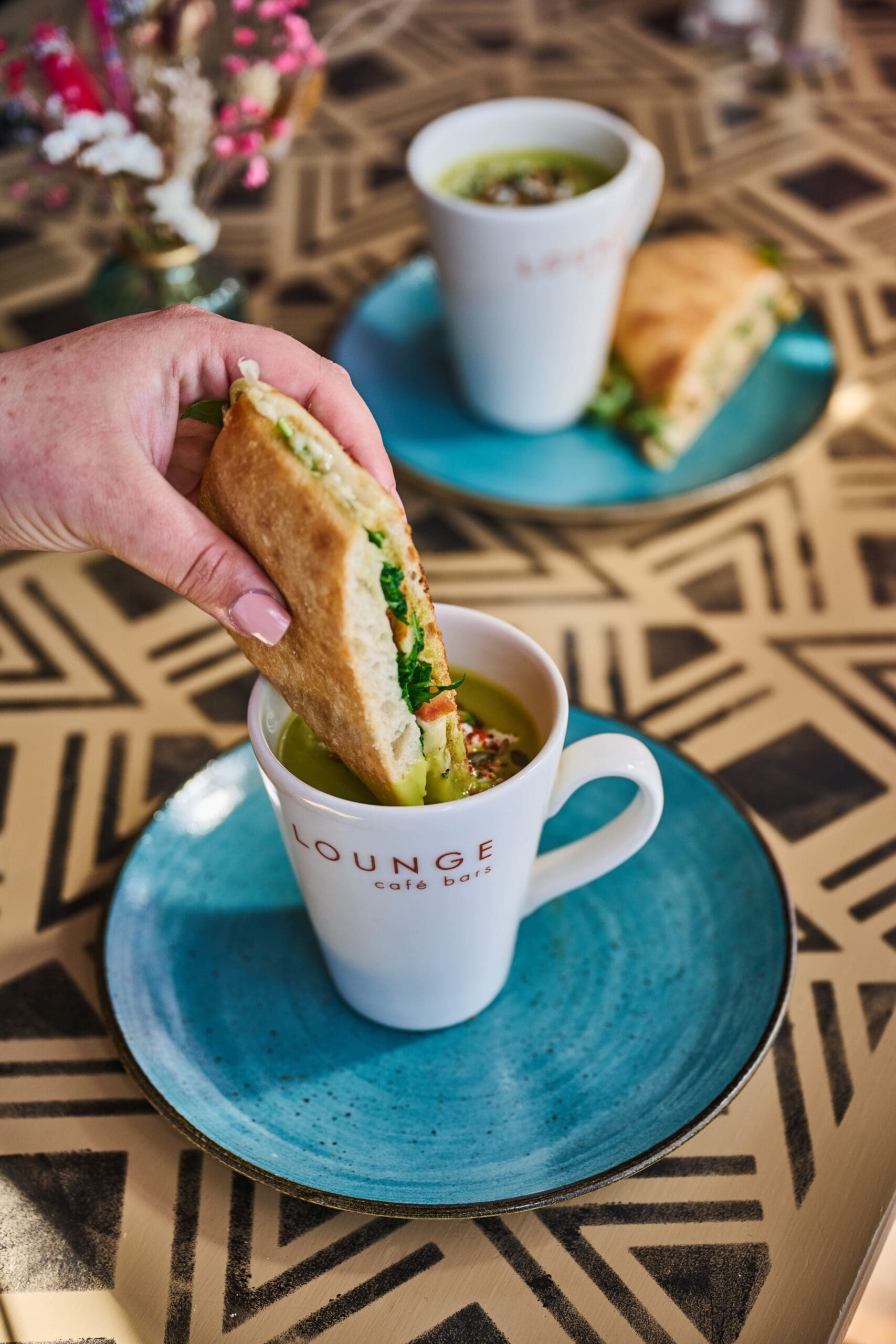 A hand dips a sandwich into a white mug of soup on a blue plate, adding comfort to the lounge setting. Another mug of soup and a sandwich rest on a patterned table in the background.
