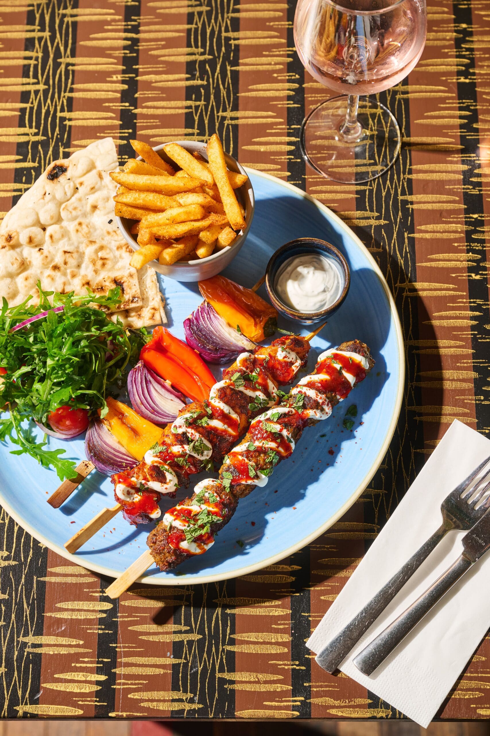 A blue plate with grilled skewers, pita bread, fries, grilled vegetables, salad, and a dipping sauce sits on a patterned lounge table next to a glass of pink drink and a napkin with a fork and knife.