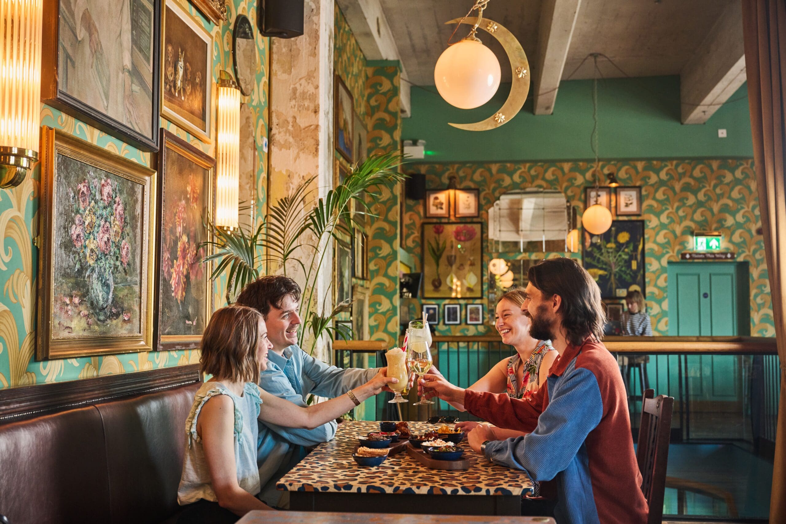 Four people sit around a table in a stylish, retro-themed lounge, raising their glasses in a toast. The decor features green and gold wallpaper, framed art, and a crescent moon-shaped light overhead.