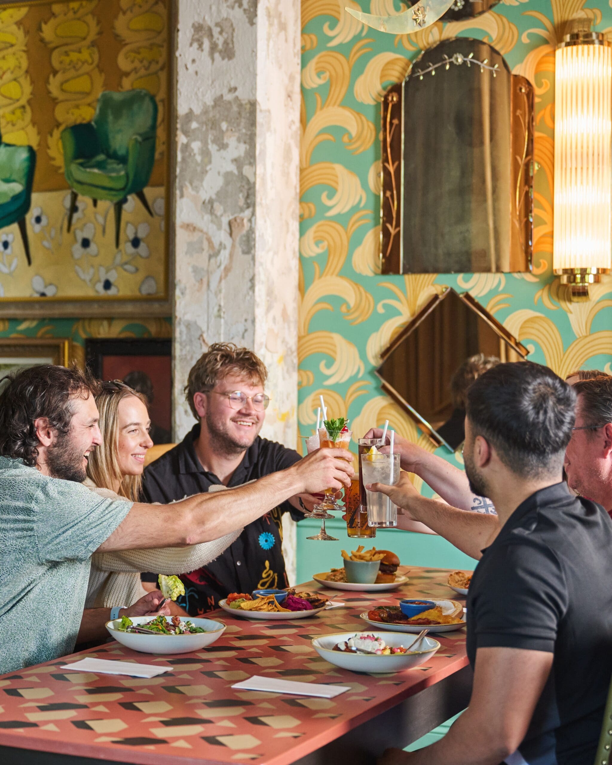 Five people sit around a table in a colorful lounge restaurant, raising their glasses in a toast and smiling. Plates of food and drinks cover the table, while patterned wallpaper and decorative mirrors brighten the background.