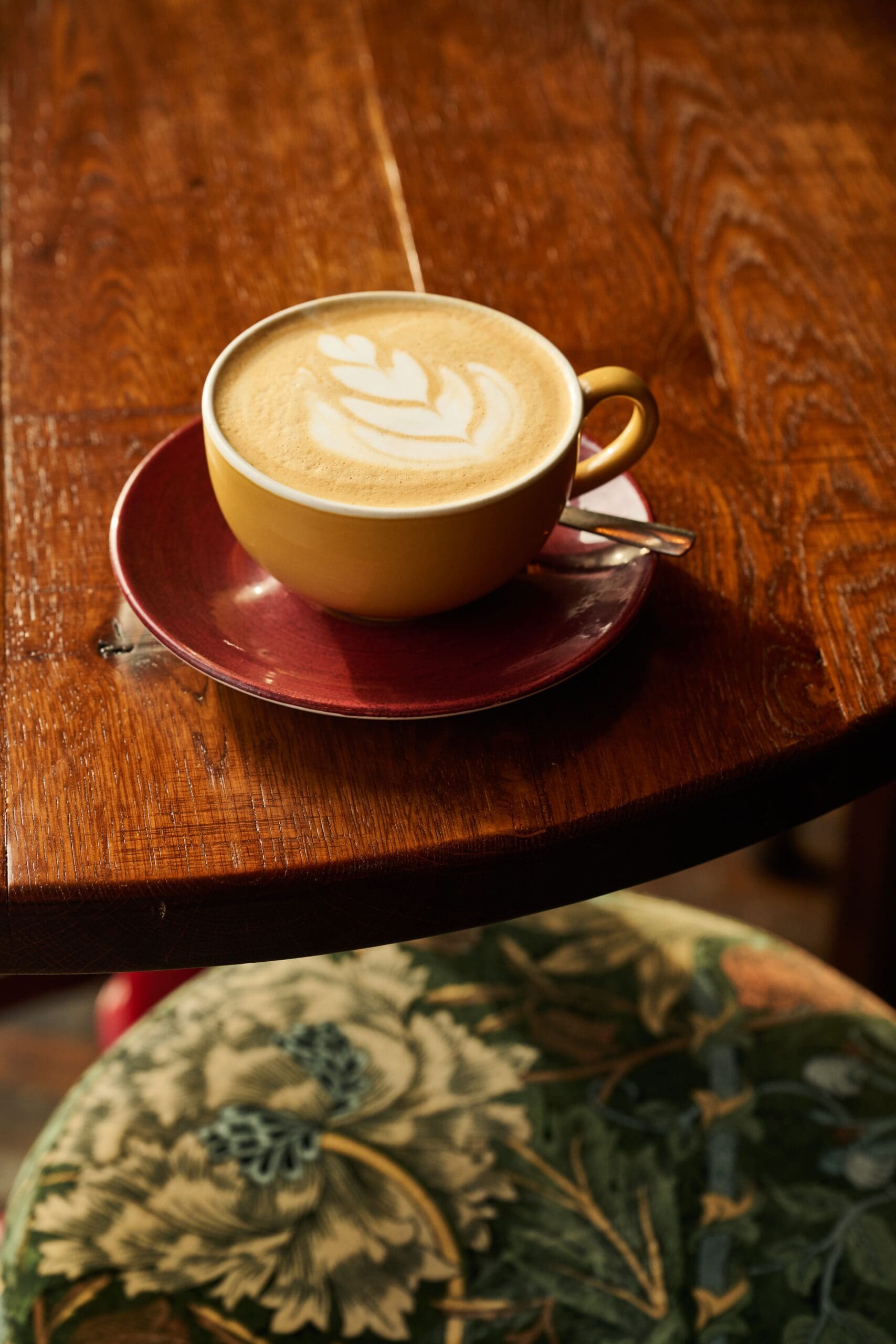 A yellow coffee cup with latte art on top sits on a red saucer with a spoon, placed on a wooden table; a green floral-patterned cushion is visible beside the table.