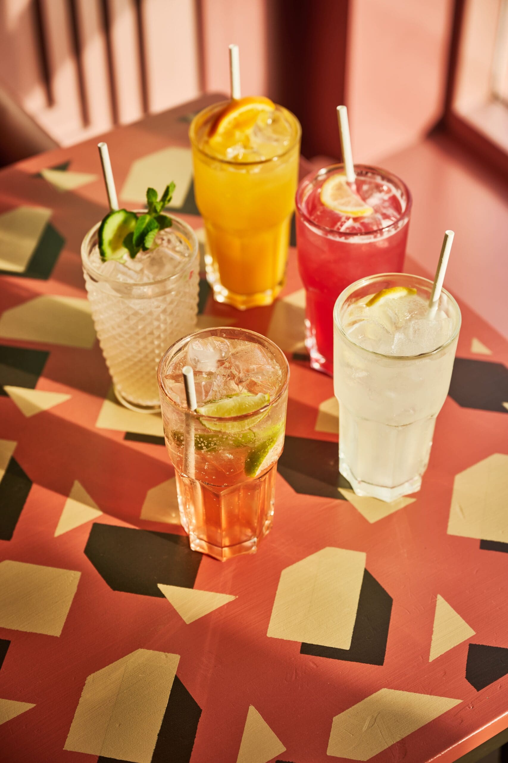 Five colorful drinks with ice and garnishes, including lime, orange, mint, and cucumber, sit on a patterned table in sunlight. Each drink has a straw.