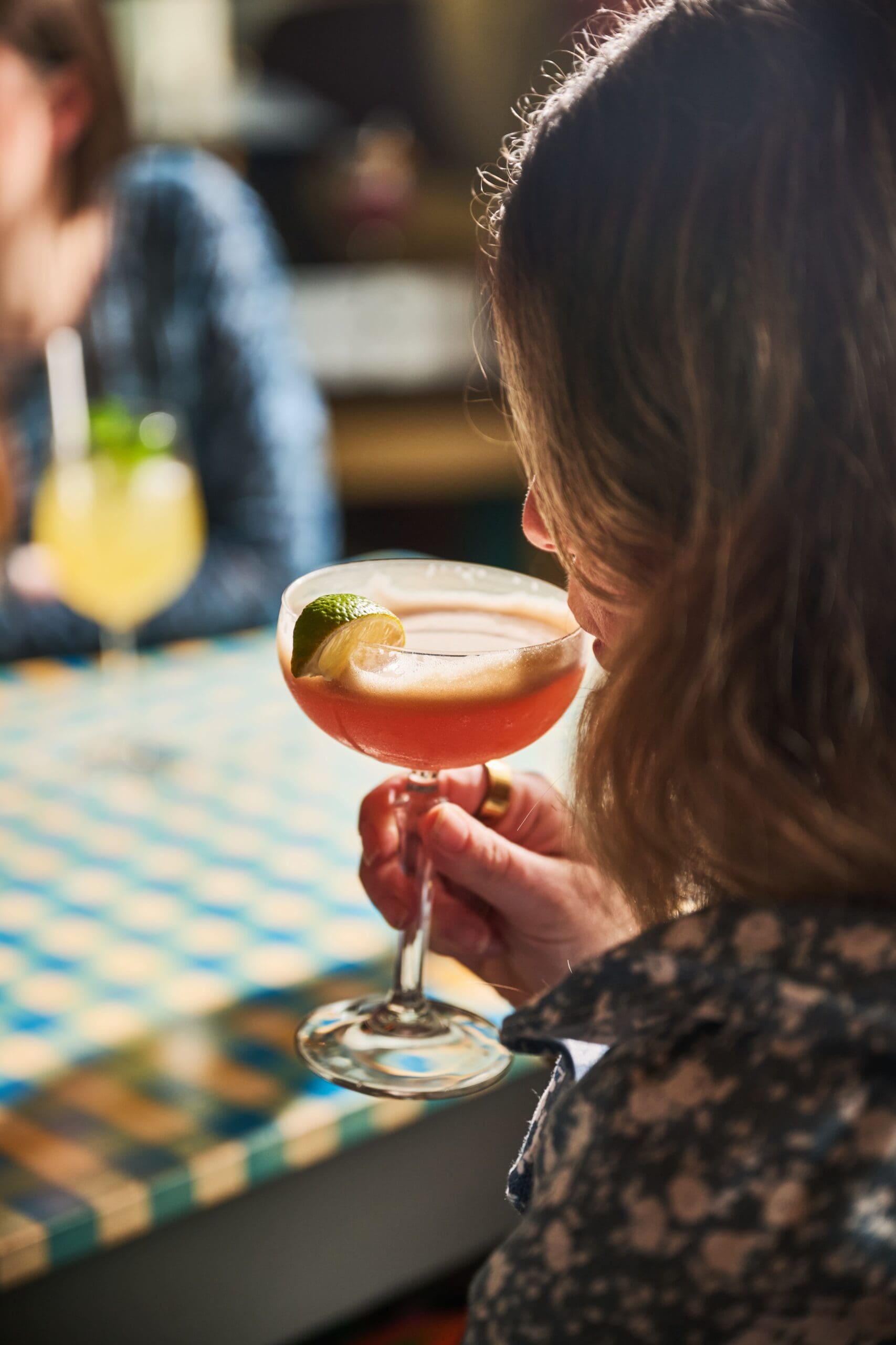 A woman holding a cocktail garnished with a lime wedge, about to take a sip, seated at a table with a checkered tablecloth. Another drink with a straw is visible in the background.