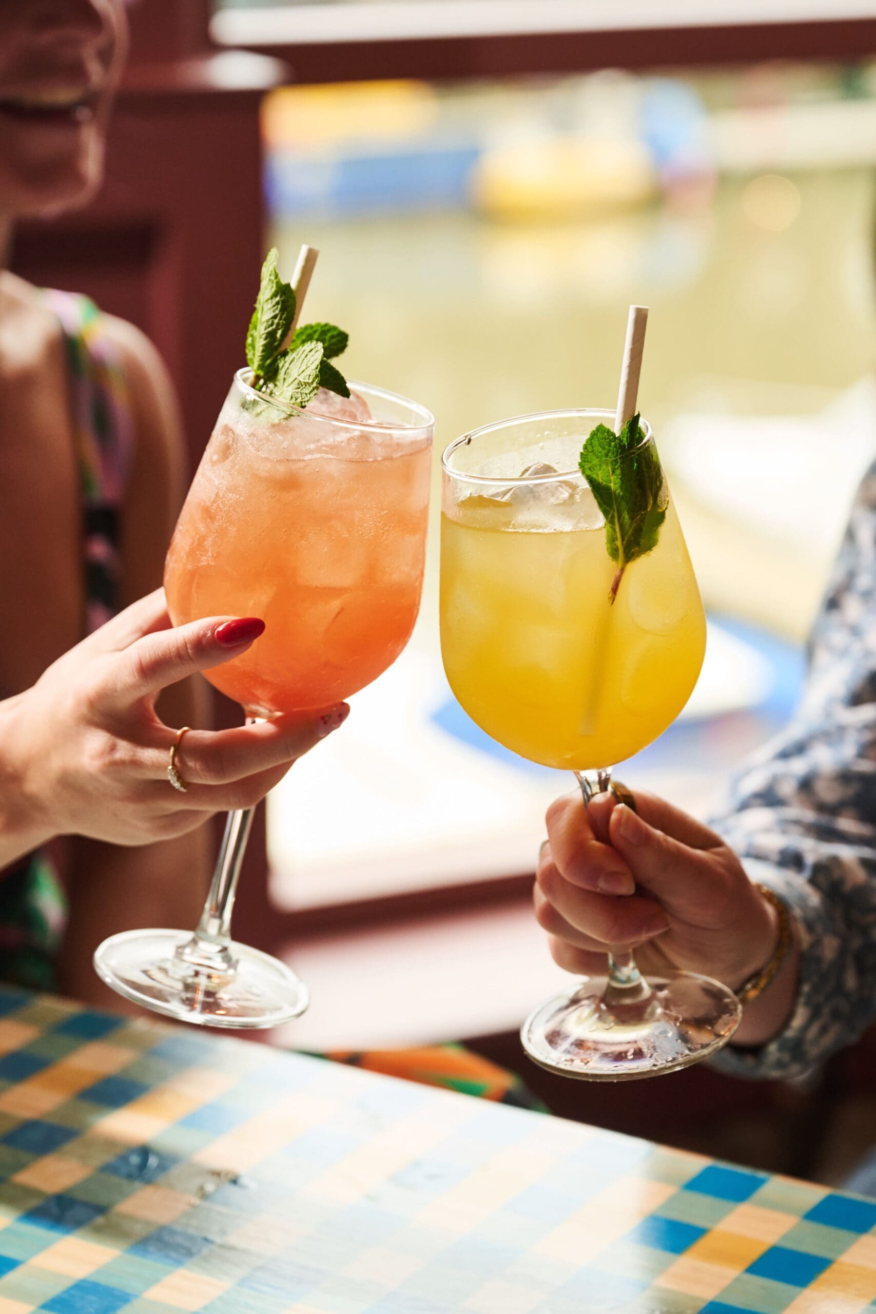 Two people clink glasses filled with colorful cocktails—one orange-red, one yellow—garnished with mint leaves and straws, as they relax at a lounge table draped in a checkered tablecloth by the window.