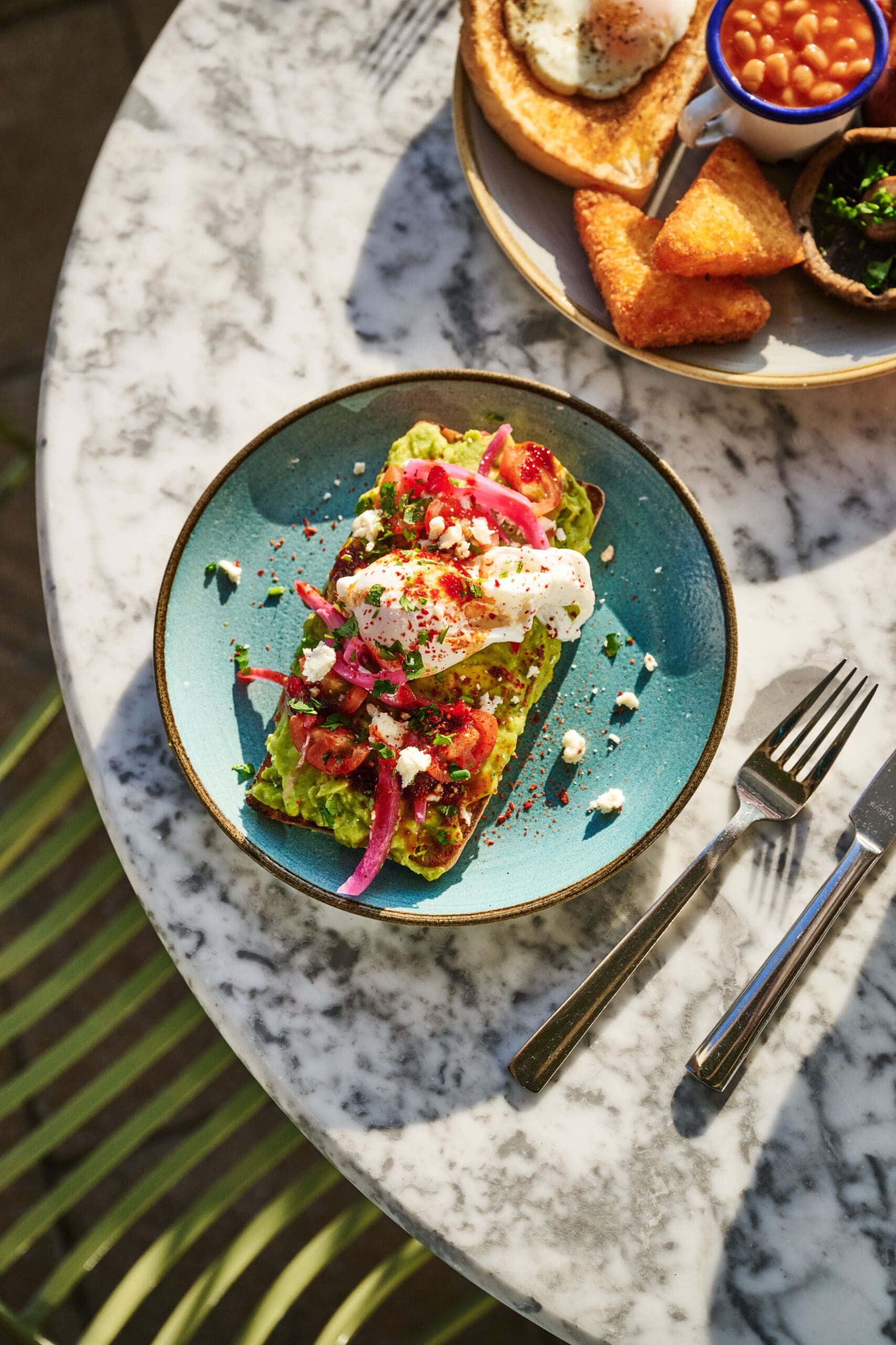 A plate with avocado toast topped with poached eggs, pickled onions, and herbs sits on a marble table, next to a fork and knife. Another plate with hash browns and beans is partially visible.