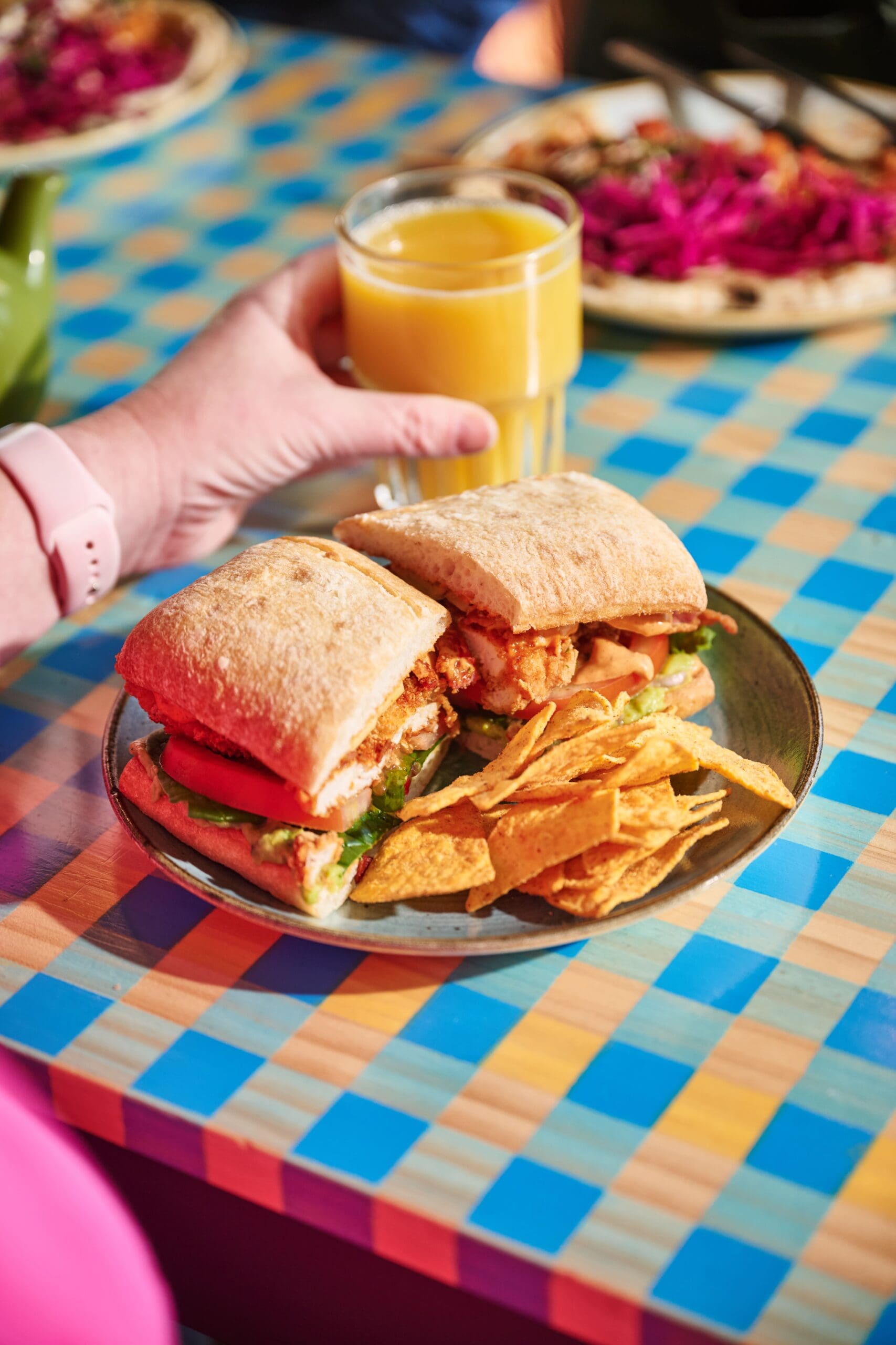 A person holding a glass of orange juice sits at a colorful checkered table with a plate of sandwiches filled with vegetables and chips.