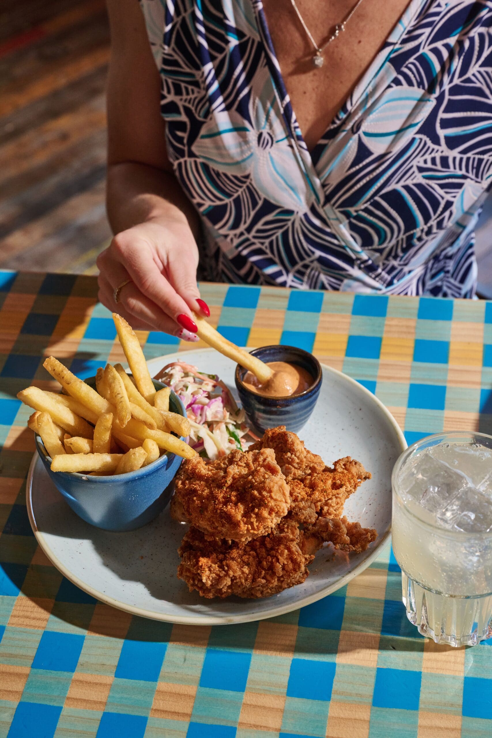 A person picks up a French fry from a plate with fried chicken, fries in a blue bowl, coleslaw, and dipping sauce. A drink with ice sits nearby on a colorful checkered table.