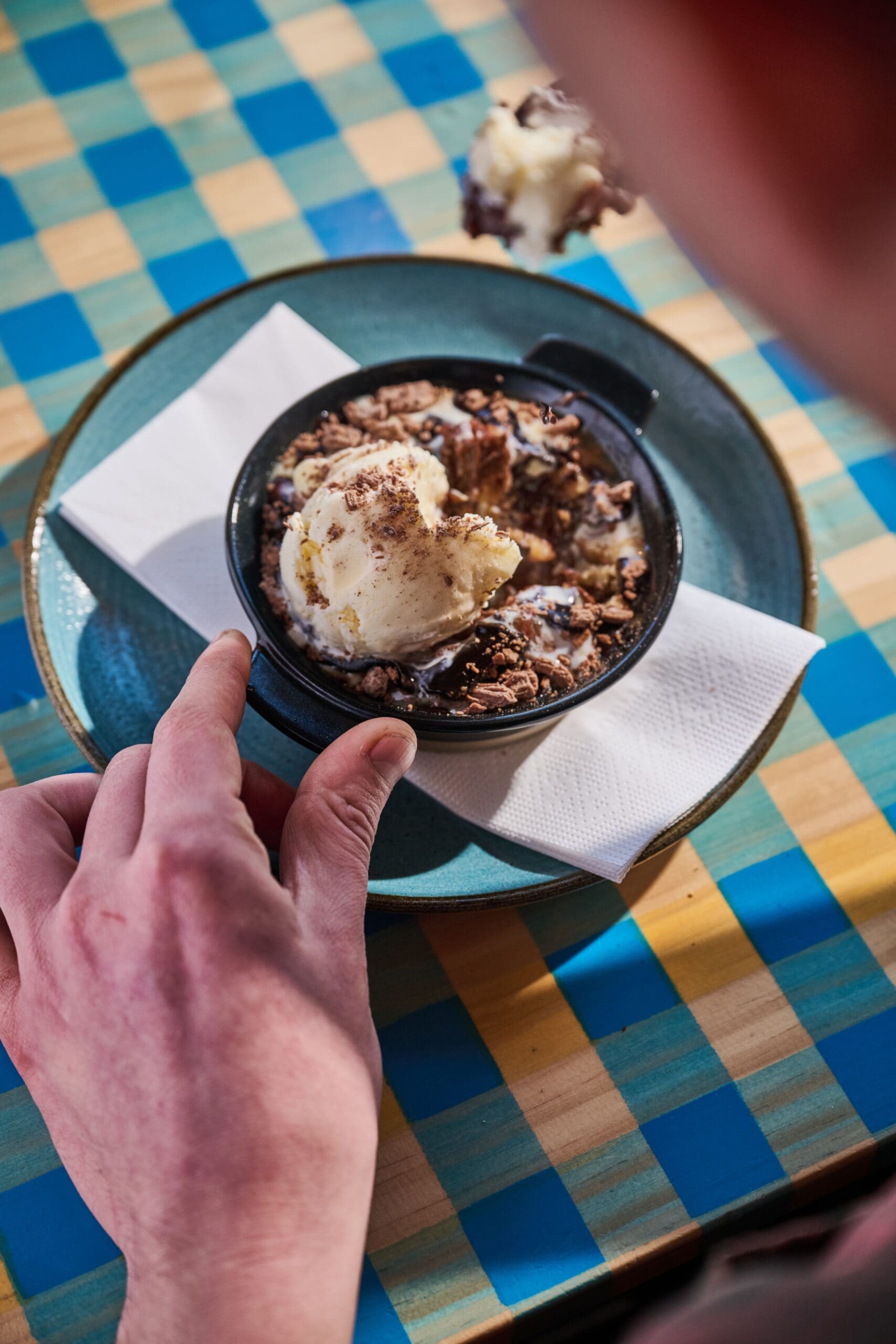 A person holds a bowl of dessert topped with a scoop of vanilla ice cream and chocolate shavings, placed on a blue plate with a napkin on a colorful checkered table.