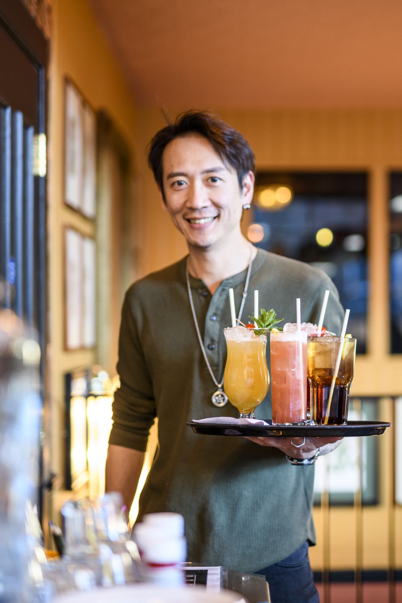 A smiling man in a green shirt holds a tray with colorful drinks, including cocktails and soda, inside a warmly lit lounge or café.