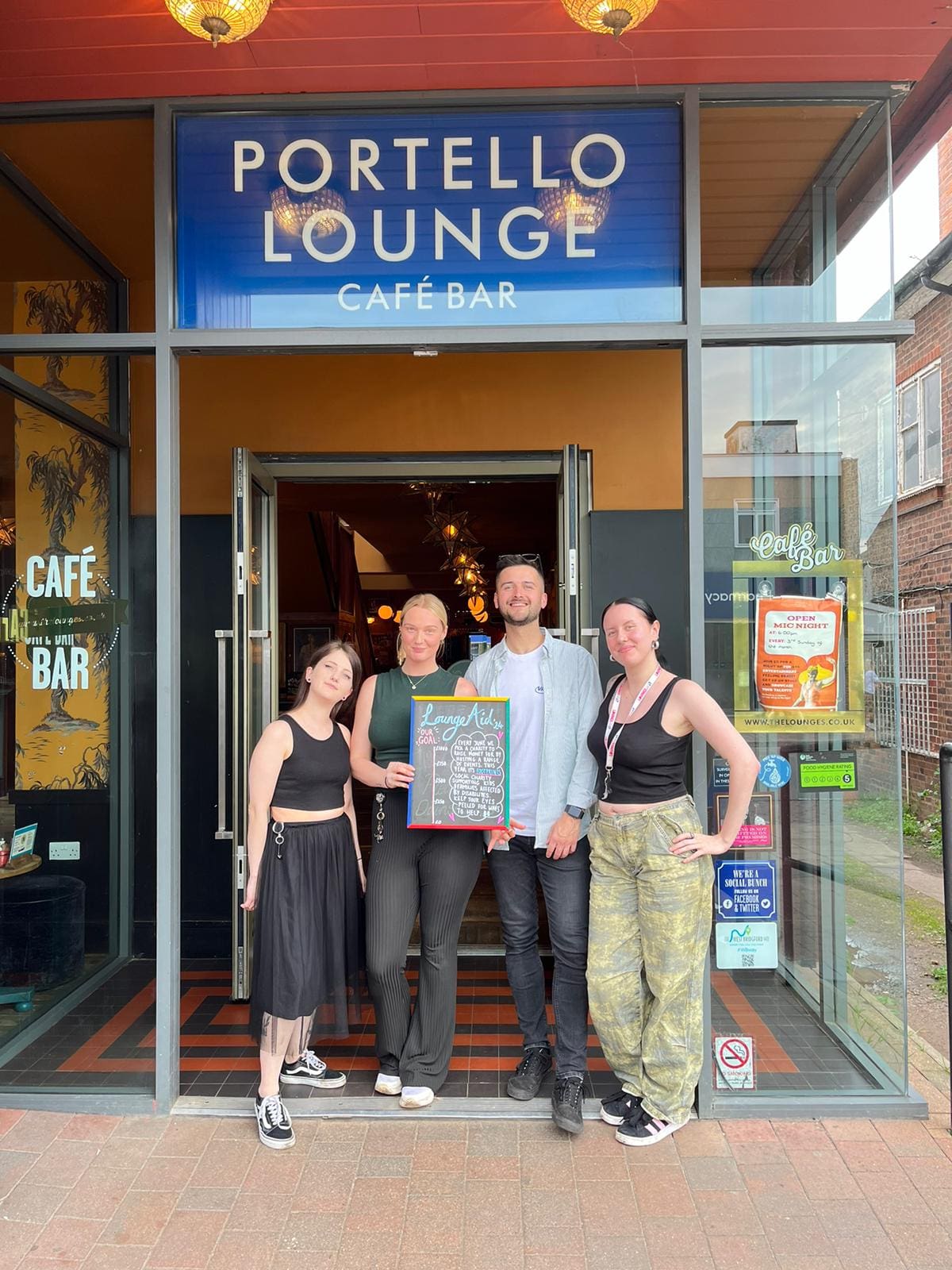 Four people stand smiling in front of the entrance to Portello Lounge Café Bar. One person holds a colorful chalkboard menu. The café sign is visible above them and bright posters are displayed at the entrance.
