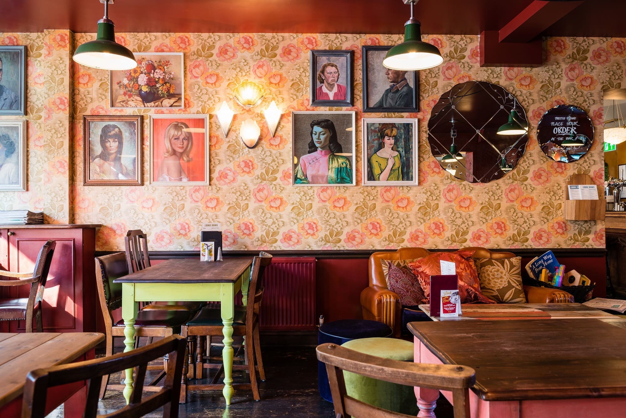 About Us: A cozy café with vintage floral wallpaper, mismatched wooden furniture, colorful cushions, and framed portraits of women on the wall, all softly lit by hanging lamps.