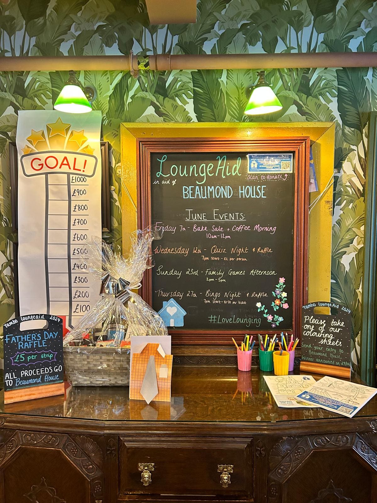 A framed chalkboard lists June events for LoungeAid at Beaumont House. Next to it, a fundraising goal thermometer, raffle display, and table with raffle tickets, colored pens, flyers, and a Father's Day raffle sign are visible.