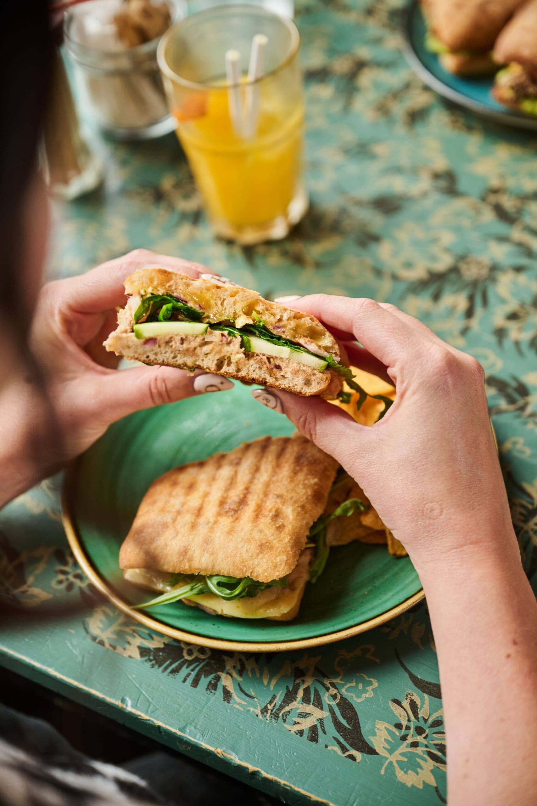 A person holds a sandwich with greens and cheese, ready to eat, while another sandwich and a glass of orange juice sit on a patterned green table.