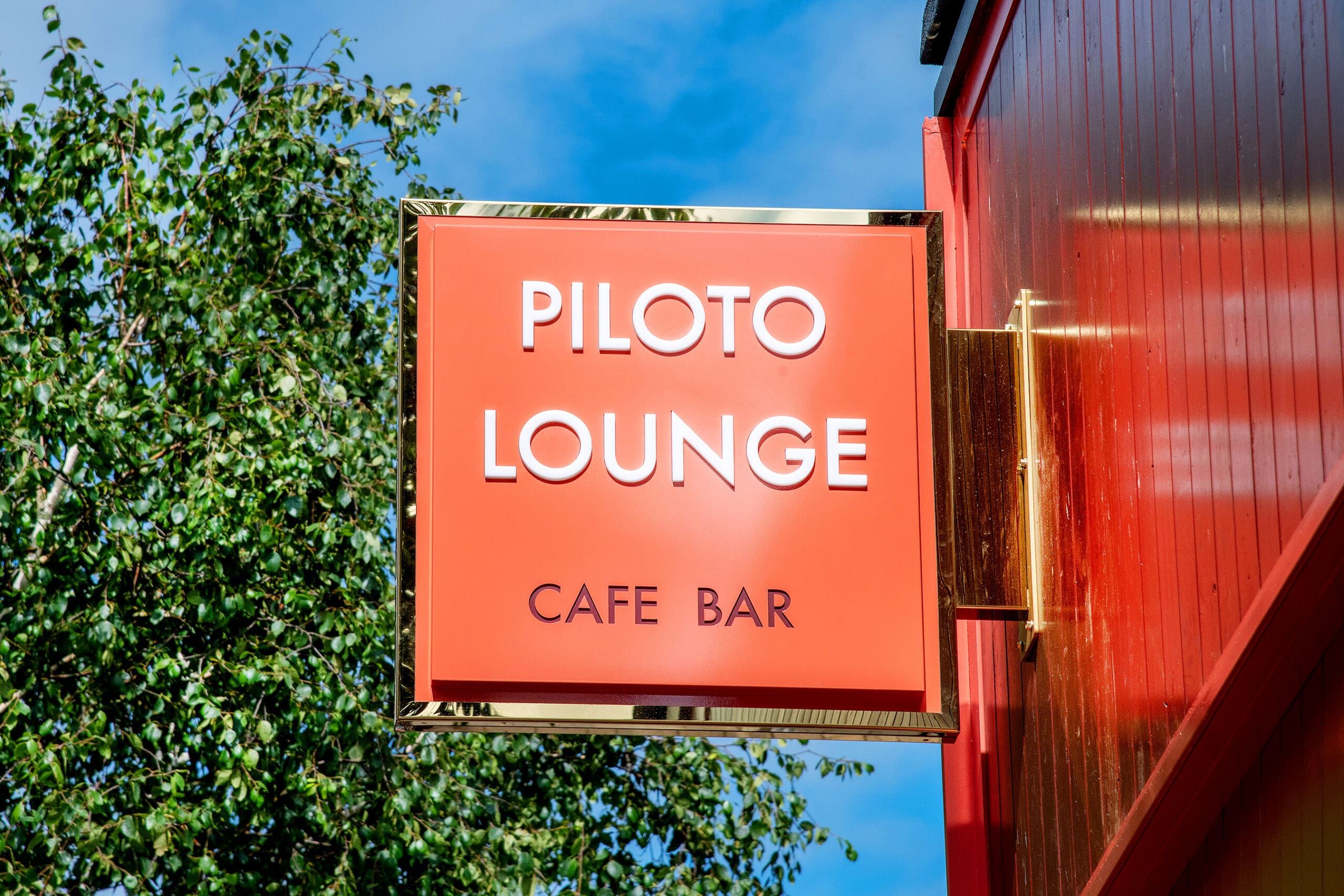 A bright red and gold sign reading "Piloto Lounge Cafe Bar" is mounted on the side of a red building, with green leafy tree branches and a blue sky in the background.