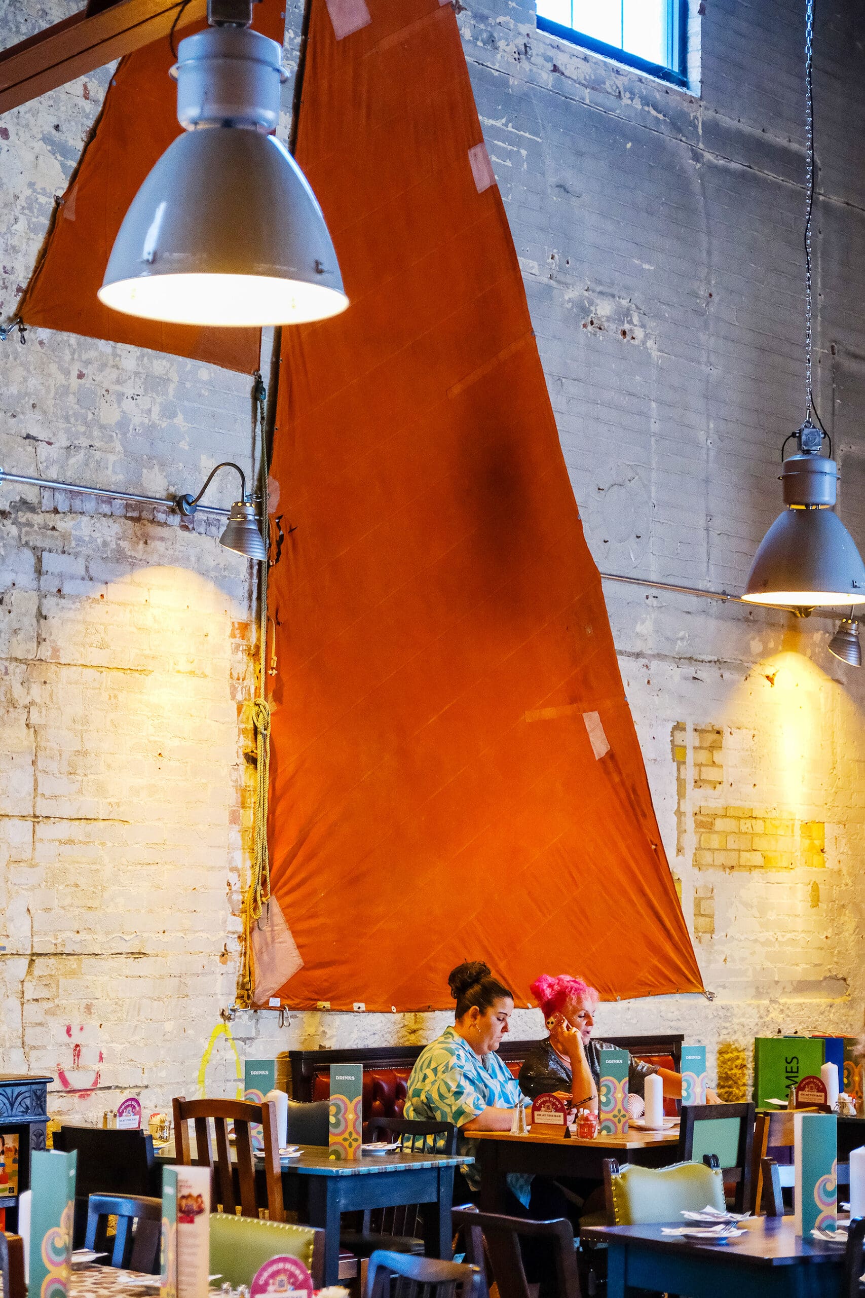 Two people sit at a table in a colorful, industrial-style cafe with exposed brick walls and large hanging lights. A striking red triangular fabric, reminiscent of a marinero's flag, hangs as decoration behind them.