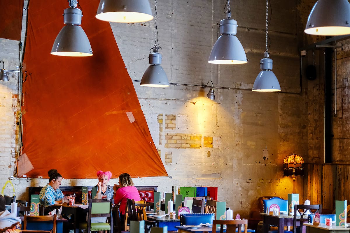 Three people sit at a table in a colorful, eclectic cafe with exposed brick walls, large orange fabric art, hanging industrial lights, and mismatched furniture. A marinero motif subtly decorates the mostly empty space around their group.