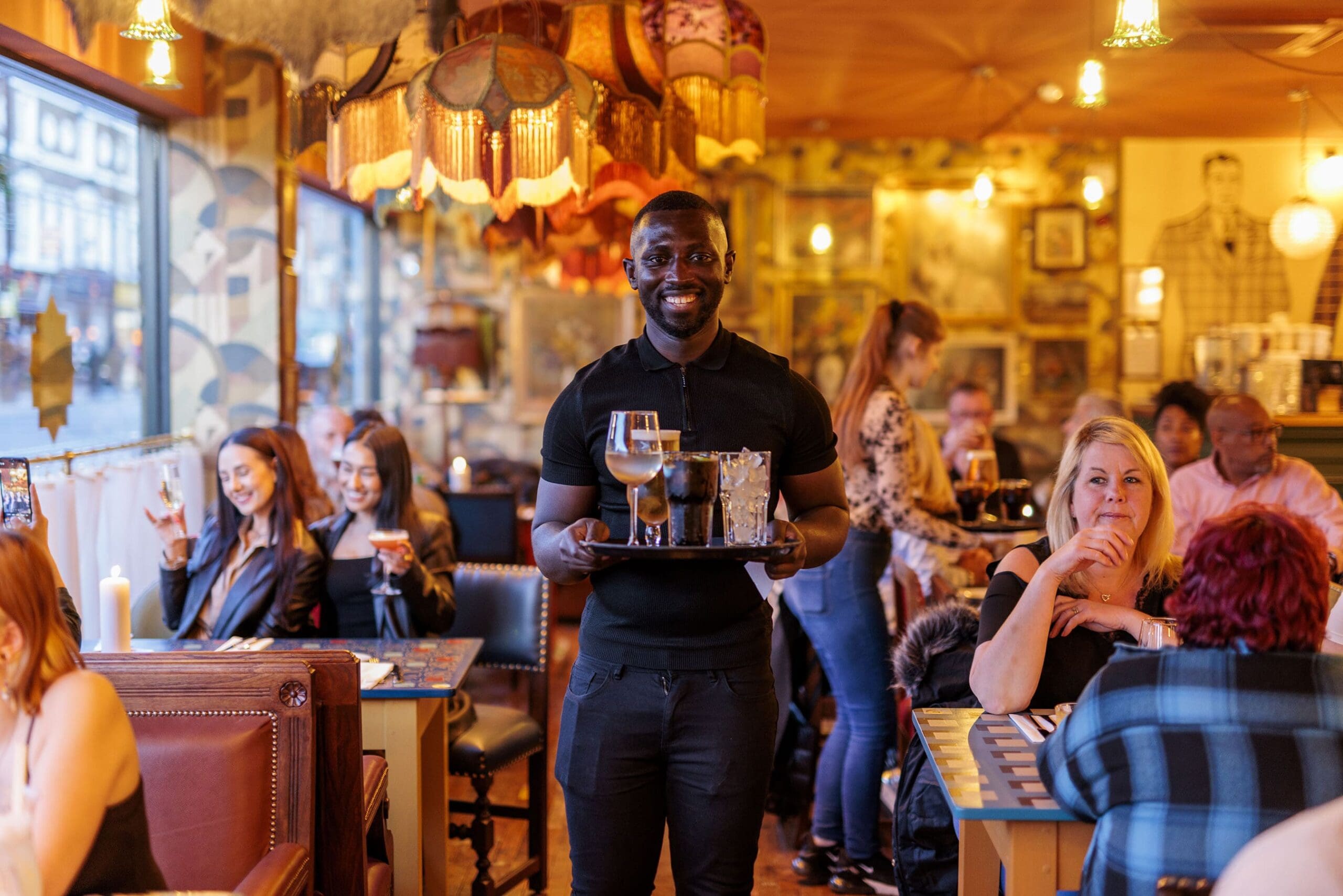 A smiling waiter holds a tray with drinks in a busy, warmly lit restaurant filled with people dining and chatting at their tables.