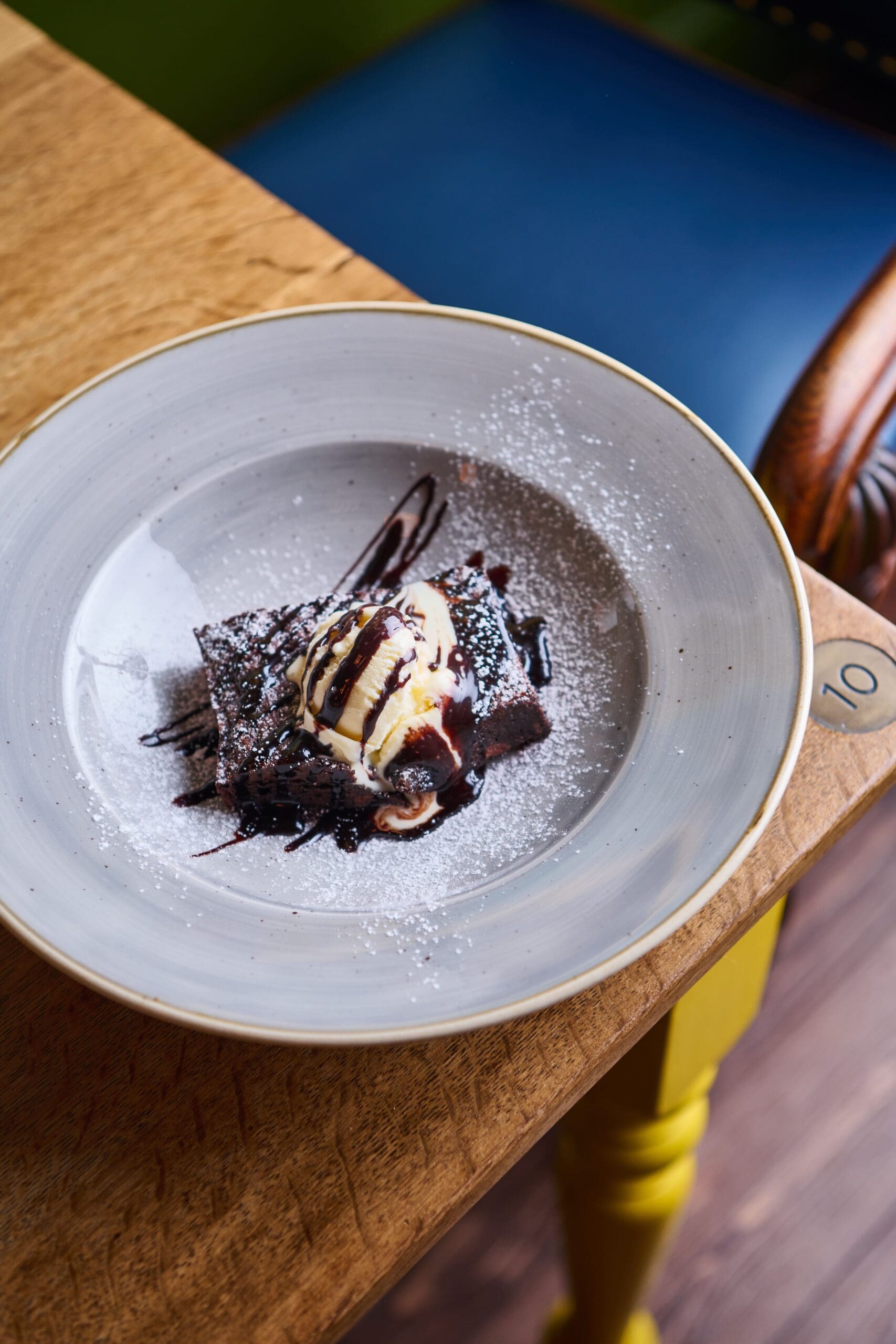 A dessert plate with a chocolate brownie topped with a scoop of vanilla ice cream, drizzled with chocolate sauce and sprinkled with powdered sugar, sits on a wooden table marked with the number 10.